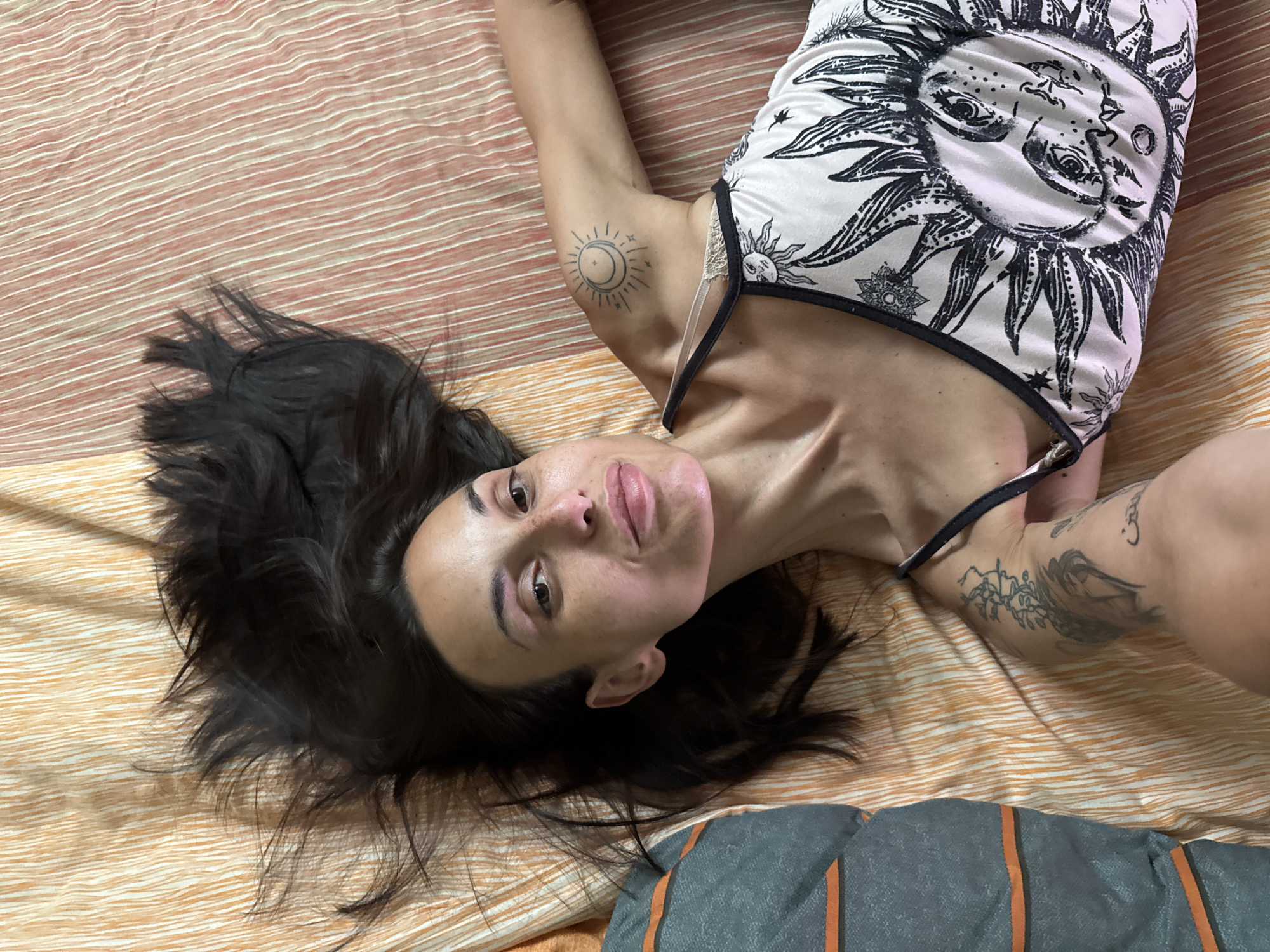 The image shows a woman lying on a bed with a striped orange and white comforter. She has long, dark hair spread out around her head. Her skin appears pale with some redness on her cheeks and nose. She is wearing a white tank top with a black trim and a large sun graphic design on it. The woman has several tattoos, including a sun and moon design on her right shoulder and what appears to be script and floral designs on her left arm. A pillow with grey and orange stripes is visible to the left of the frame. The scene is a close-up, taken from a high angle. The exact location within Pleven, Bulgaria, cannot be determined from the image.