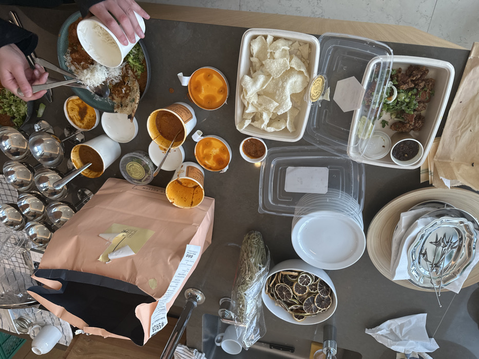 An overhead view depicts a kitchen counter laden with food and serving items, indicating a meal in progress, likely takeout. The location is identifiable by a label on a large brown paper bag, which reads "OLD ST. BOTOLPH," a reference to an area in the City of London, United Kingdom.
At the upper right, two hands are visible: one uses a serving spoon to transfer white rice from a small white paper container onto a large blue-green plate, while the other hand holds the rice container. The large plate also contains a dark orange curry, what appears to be naan bread, and green garnishes.
Spread across the dark grey countertop are numerous food items and containers. These include:
* Multiple white paper cups, some partially emptied, containing orange-brown curry or sauce, with spoons present in some. Two wider, white paper containers are also present, possibly for additional sauces or soup.
* A rectangular paper container holds numerous light-colored, wavy crackers (pappadums).
* Another rectangular paper container contains fried protein pieces, garnished with green herbs and possibly a lime wedge, accompanied by small plastic cups of dark and white sauces.
* A small clear plastic container with a green substance.
* A small plastic cup with red-orange sauce.
* A white bowl filled with dried citrus slices.
* A clear plastic bag containing a bundle of thin, green, dried strands, possibly herbs or noodles.
* A stack of small, white ceramic plates.
* A wooden serving platter holds several silver-colored, scalloped-edge plates and silver serving tongs.
* A rack at the top left displays multiple small, reflective silver bowls.
* A white ceramic jug and a single white mug are visible near a metallic faucet and sink area on the left.
* Several clear plastic container lids are also present on the counter.
The overall scene conveys a casual dining setup with an emphasis on shared portions from delivered food.