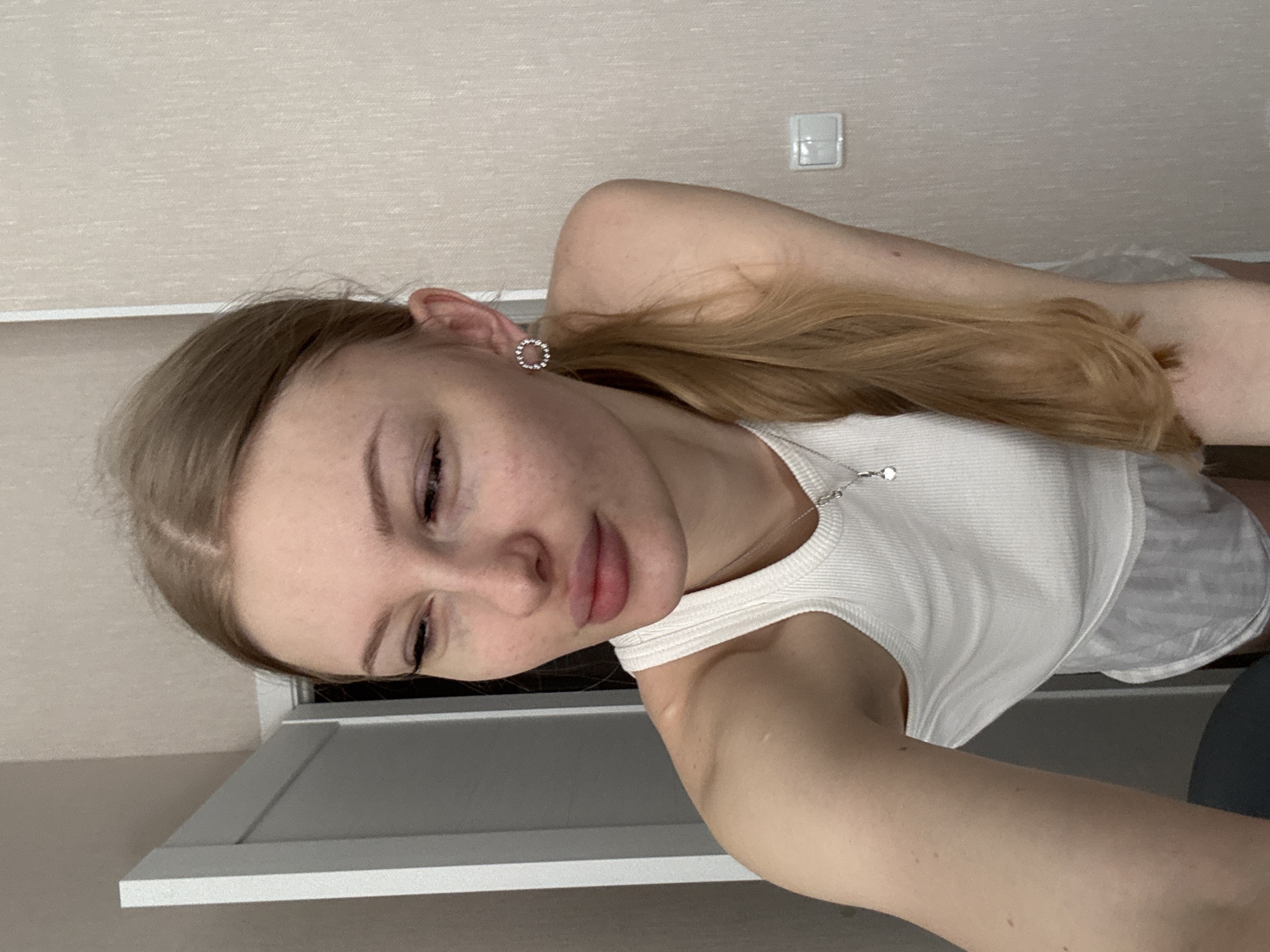 A woman is depicted from a high angle. She is wearing a white tank top and appears to be indoors. She is fair skinned, with light brown hair pulled into a ponytail. She is wearing a circular earring in her left ear and a necklace with a small pendant. The background includes a beige wall and part of a white door frame. The location is likely Perm, Russia.