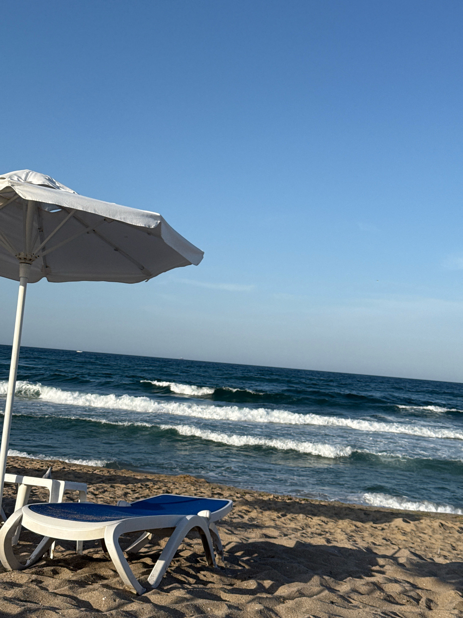 A beach scene in Sozopol, Bulgaria, features a white beach umbrella casting a shadow on the sand. In the foreground, a white and blue plastic sun lounger rests on the sand, angled towards the sea. The ocean is visible in the background with clear blue water and white waves. The sky is a bright, cloudless blue. The overall impression is a tranquil beach setting.