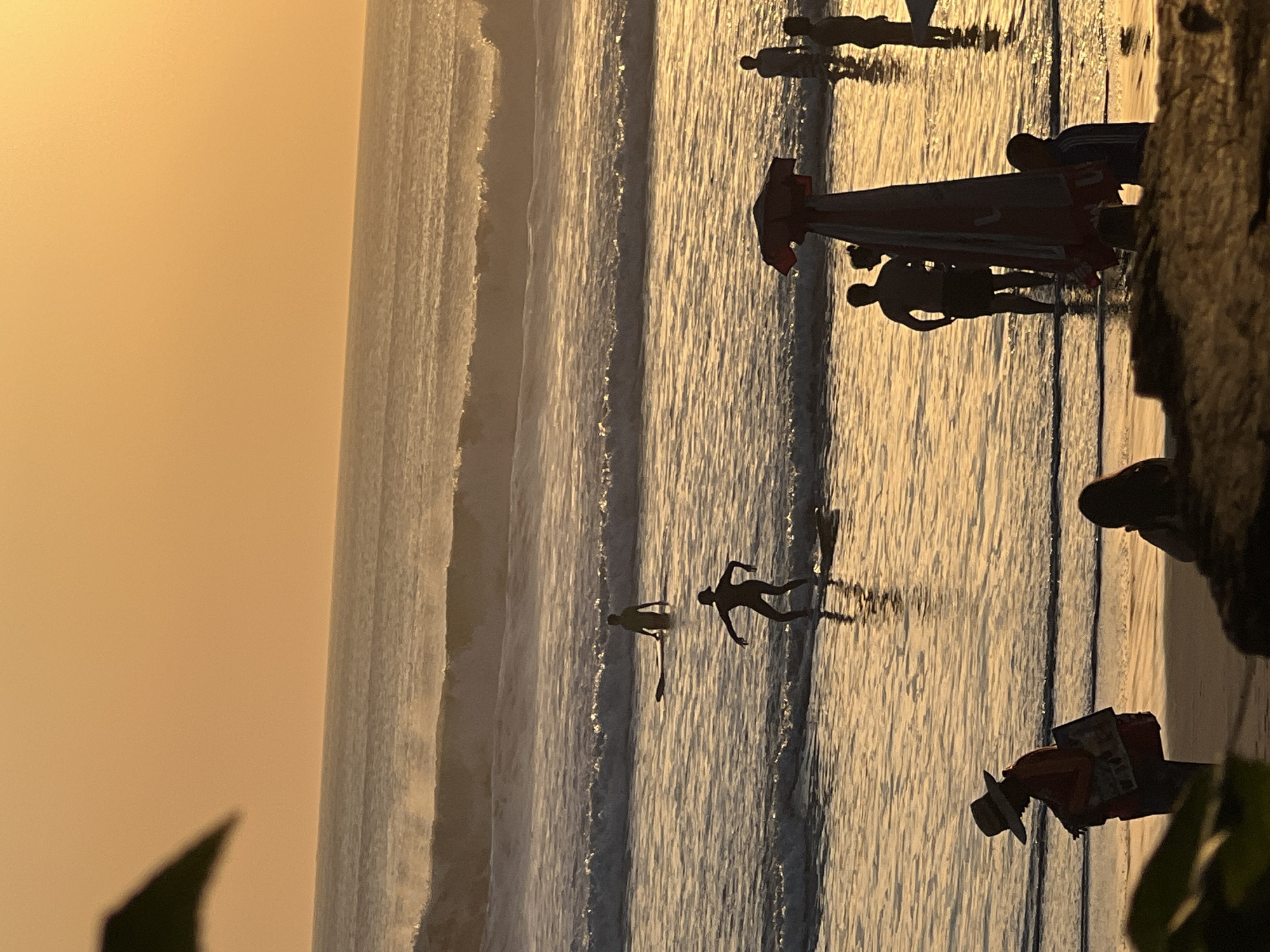 The image depicts a beach scene at sunset in Kuta, Indonesia, characterized by the warm, orange glow of the setting sun reflecting on the water. Several individuals are present. In the foreground, silhouetted figures are visible on the beach. One individual, wearing a wide-brimmed hat, stands facing left, holding a bag. To their right, another person is seated. In the middle ground, two people are surfing; one is actively riding a wave, while another stands on their board in shallow water. Further out, another surfer is visible in the distance. The ocean features moderate waves breaking near the shore, with multiple sets of waves visible extending towards the horizon. A beach umbrella is partially visible in the mid-foreground. The overall atmosphere is serene, with the silhouettes and warm lighting suggesting a peaceful end to the day.