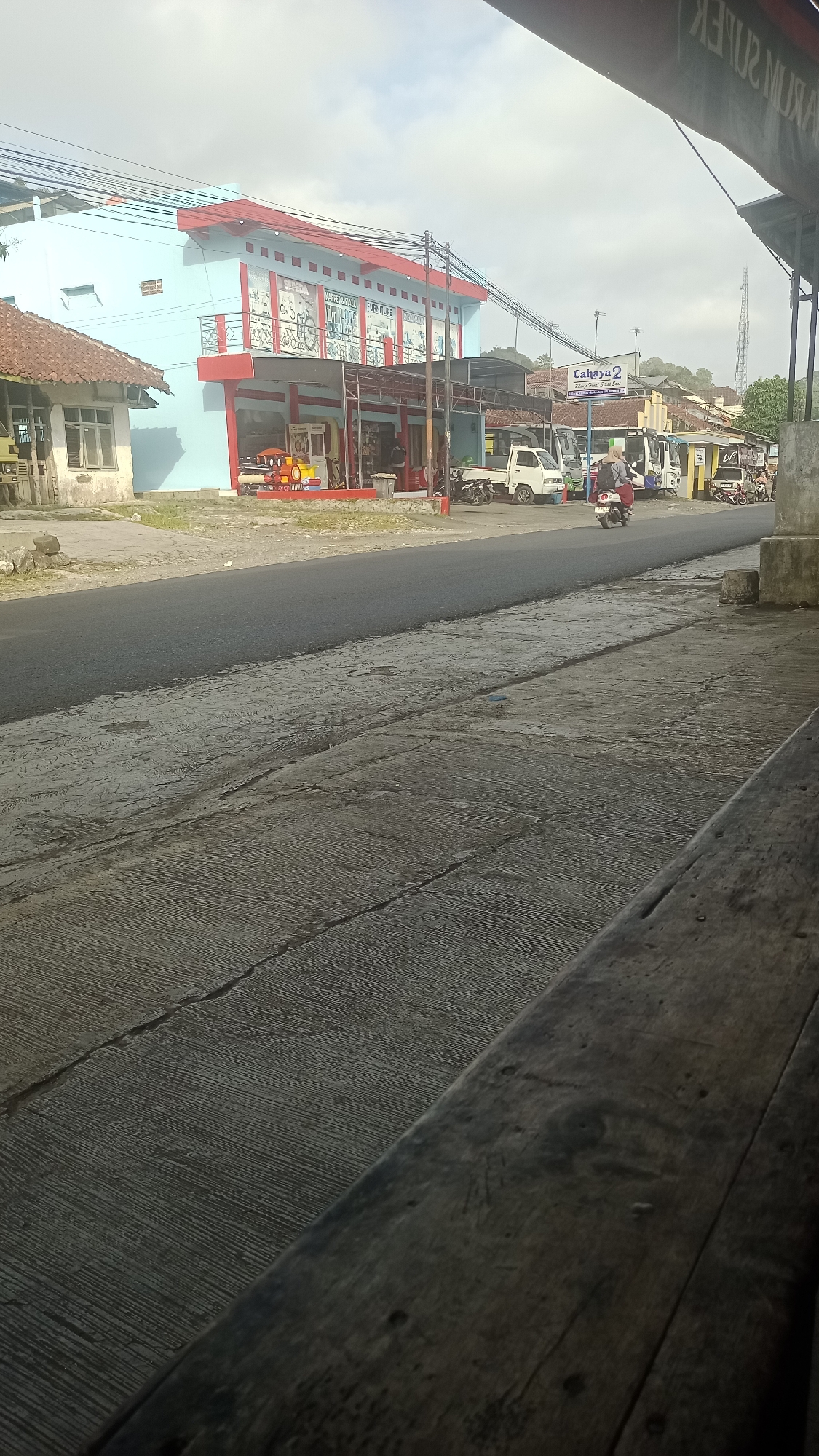 The image shows a street scene in Karyamandala, Indonesia. On the left, a light blue building with a red roof and accents has signage that includes "FURNITURE". In front of the building, there are storefronts with various items displayed. A yellow and red toy ride is visible. A white truck and a few motorcycles are parked on the side of the road. A person is riding a scooter down the street. The road surface is asphalt, and the sidewalk is made of concrete with visible cracks. The sky is overcast. A sign with "Cahaya 2" is visible on a building further down the street.