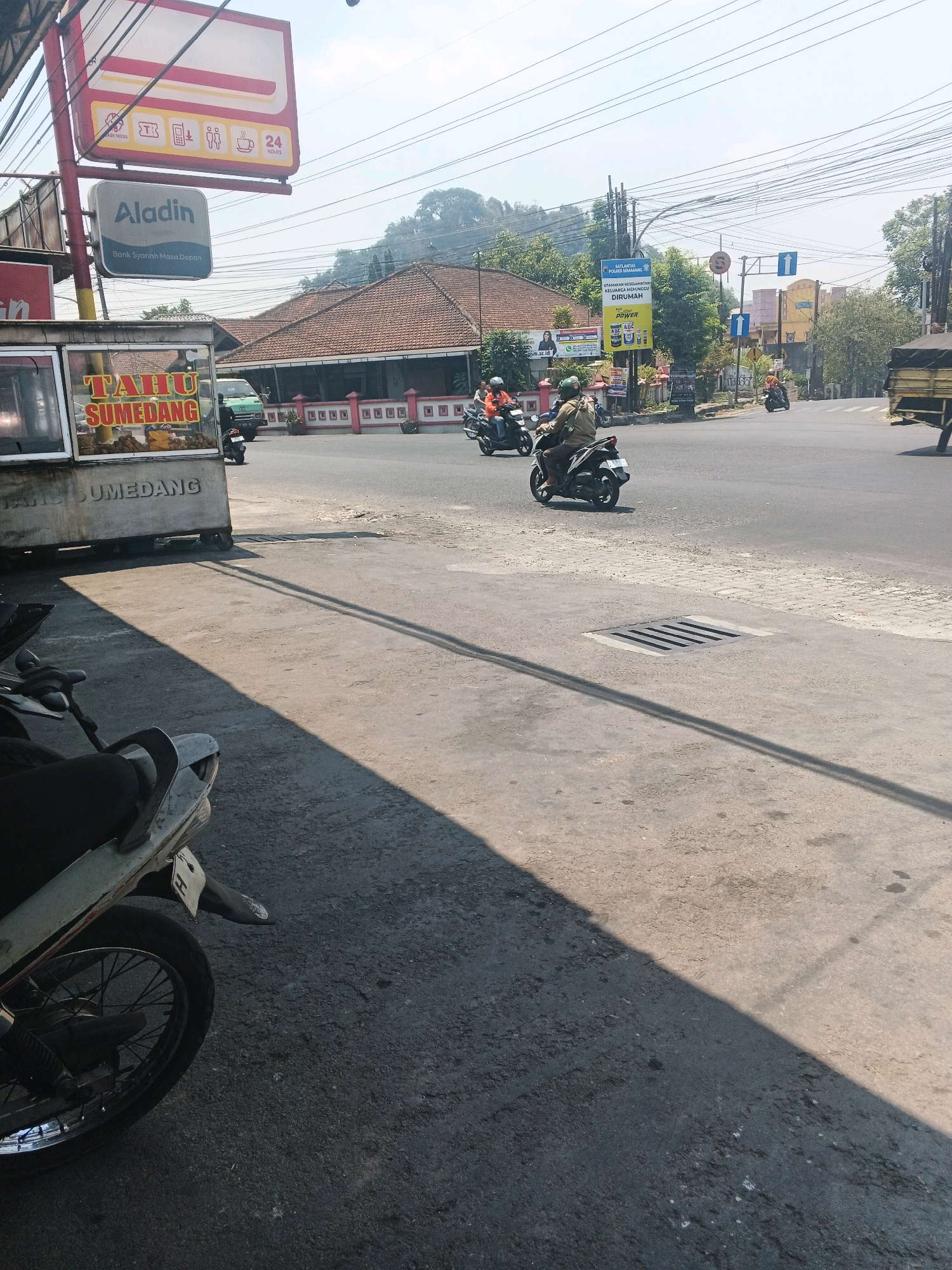 The image depicts a street scene in Ungaran, Indonesia, on a bright, sunny day. In the foreground, a motorcycle is partially visible, casting a long shadow across the asphalt. A food cart with the words "TAHU SUMEDANG" (Sumedang Tofu) emblazoned on its side is situated to the left. In the mid-ground, several motorcycles with riders are navigating a road intersection. One rider is wearing a brown jacket and a helmet, while another, on a red and white motorcycle, is wearing an orange shirt and helmet. A green truck is parked in the background, and a building with a terracotta-tiled roof is visible behind it. A large billboard with an advertisement is also present. The sky is clear with a few thin clouds. Overhead power lines are a prominent feature of the environment.