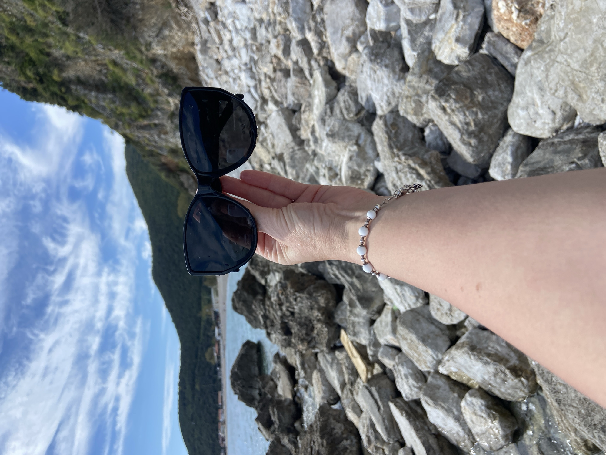 A right hand wearing a bracelet with alternating white and silver beads is holding sunglasses in front of a rocky shoreline in Prijevor, Montenegro. The hand is tanned, and the bracelet is fastened near the wrist. The sunglasses have black frames and dark lenses. In the background, a body of water is visible with a coastline and a hillside with vegetation under a blue sky with clouds. Rocks and pebbles form the ground surface under the hand. The image is slightly rotated.