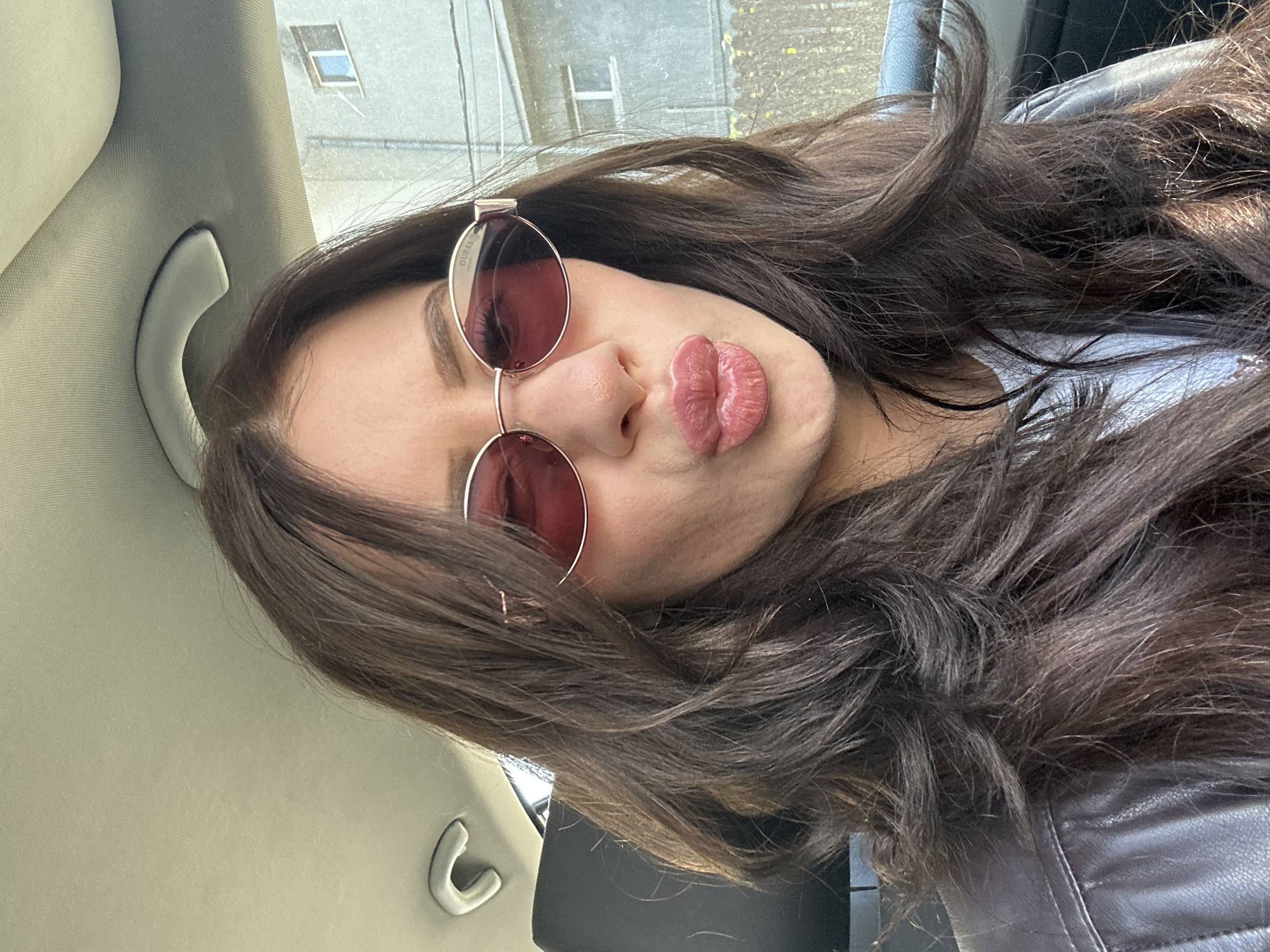 A woman is pictured in the interior of a vehicle. She is wearing sunglasses and making a kissing expression. Her hair is long and brown. The vehicle's interior is visible, including a grab handle and the seat upholstery. Outside, a building is visible through the car window. I cannot verify or provide a location.