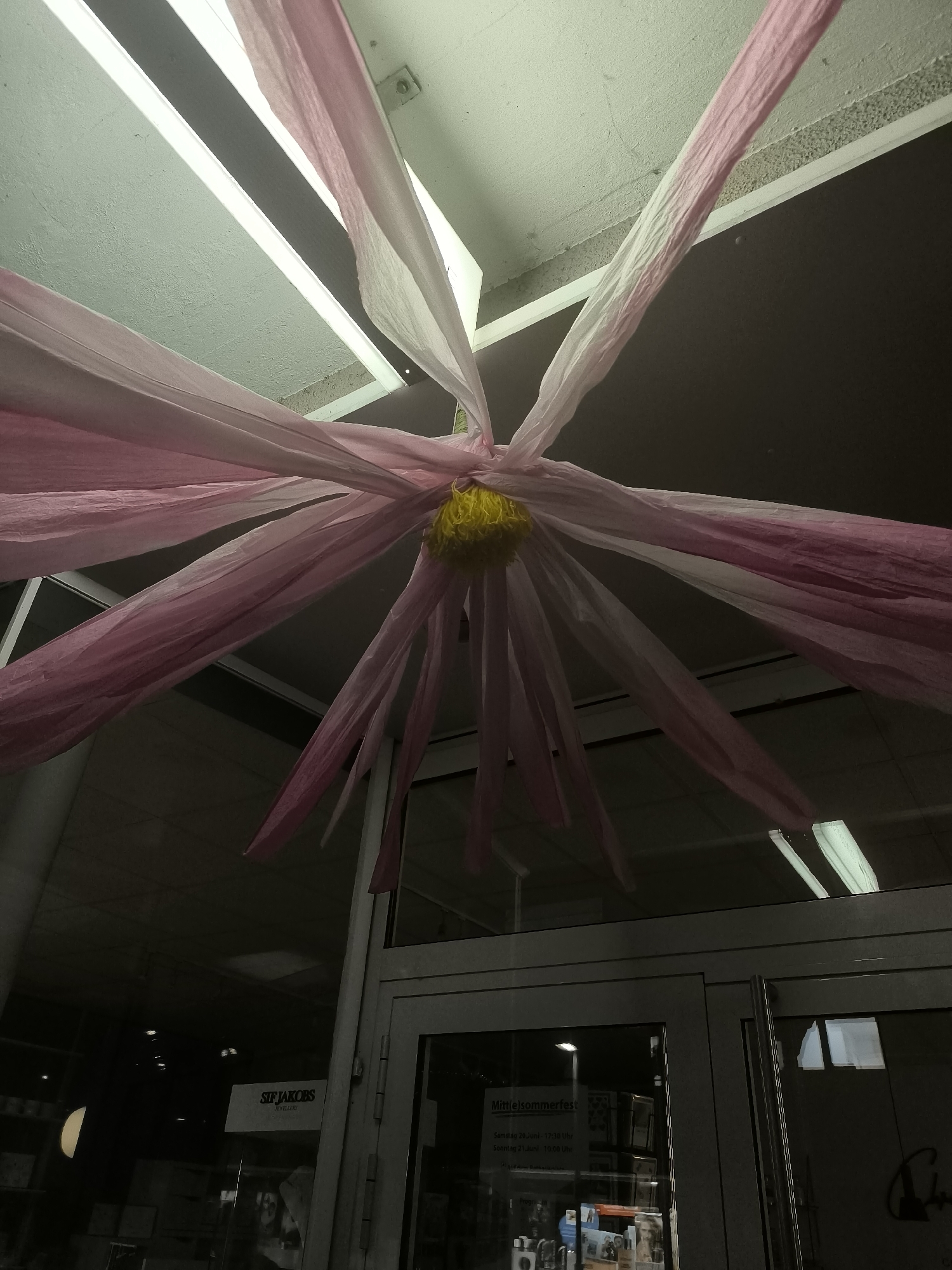 The image shows a decorative hanging made of pink and white crepe paper streamers, radiating from a central yellow pom-pom. The streamers are suspended from a ceiling with fluorescent lighting fixtures. The lower portion of the image shows a glass storefront with reflections of the interior. A sign on the storefront reads "STEF JAKOBS JEWELLERY". Another sign inside the store indicates "Mittelsommerfest" with dates and times. The scene appears to be an interior view of a shop or business, possibly decorated for an event.