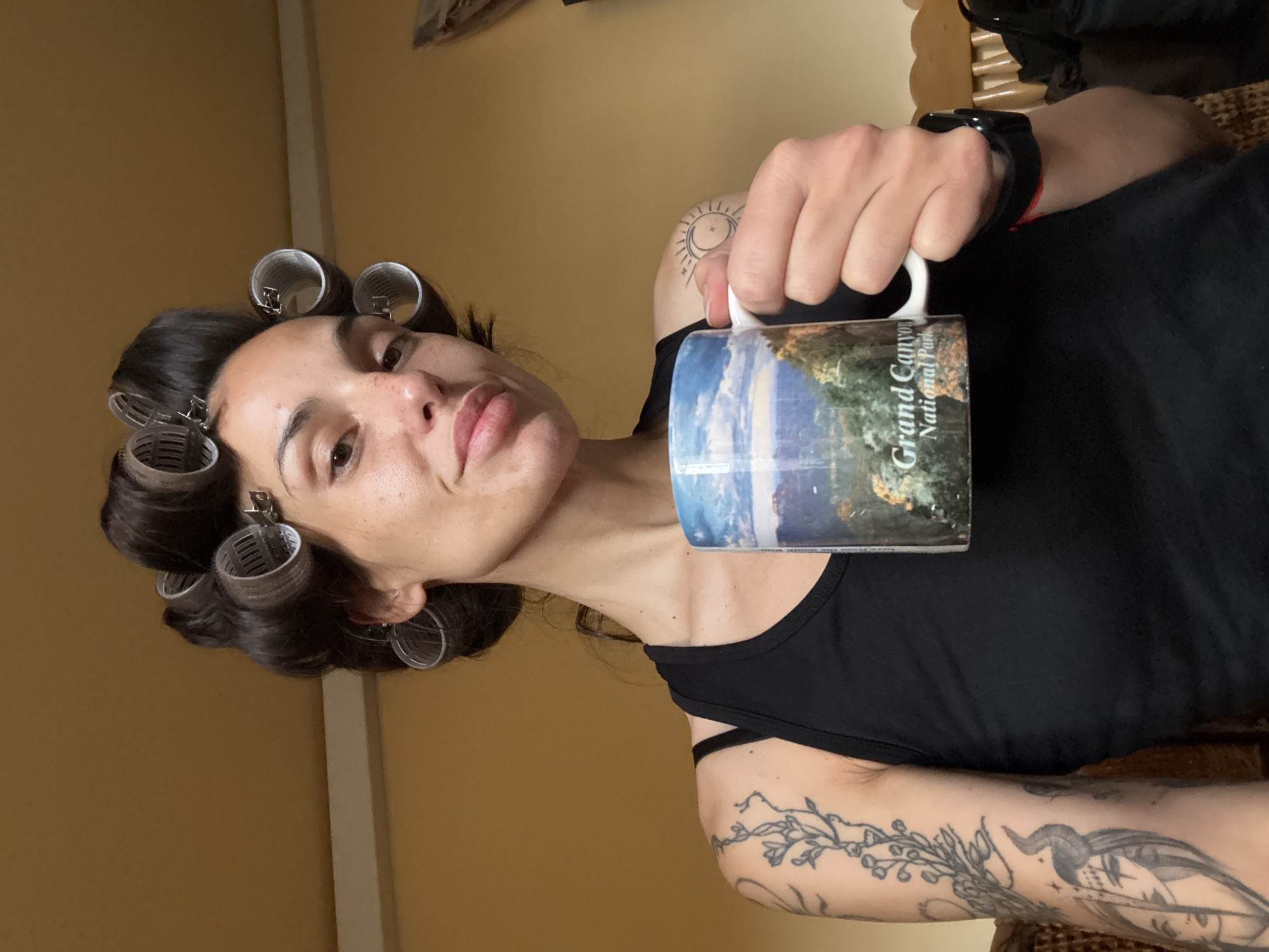 A fair-skinned person with hair curlers poses indoors, holding a "Grand Canyon National Park" mug. The person is wearing a black tank top, a black watch with a red band, and has arm tattoos. The background consists of a yellow wall and partial view of furniture. The location is not identifiable as Pleven, Bulgaria from the visual information.