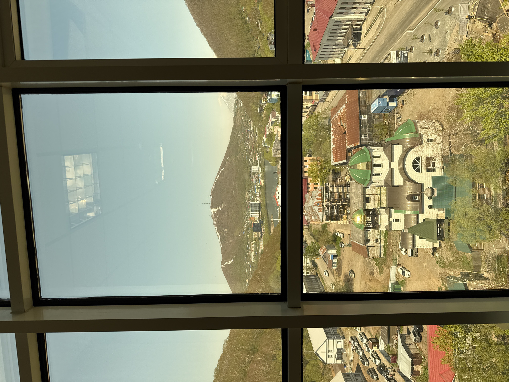 The image displays a detailed view of Petropavlovsk-Kamchatsky, Russia, captured through a multi-pane window. The scene is divided horizontally by a window frame, separating the upper sky and distant mountains from the lower urban landscape, and vertically by three additional window frames.
In the upper section, a clear, light-blue sky dominates, with a mountainous backdrop. A prominent, partially snow-capped mountain (likely Mishennaya Sopka) stands in the center-left distance, topped with several communication towers or structures. Urban development, including buildings and a body of water (Avacha Bay or a lake), extends along the base of the mountains. Faint reflections of interior overhead grid lighting are visible on the window glass in the upper left and right quadrants.
The lower section provides a close-up of the city's architecture and street life. A distinct white Russian Orthodox church with multiple green, onion-shaped domes and a prominent arched entrance is centered in the mid-ground. Surrounding the church are diverse buildings, including older multi-story structures with weathered wooden or corrugated metal roofs, and newer multi-story buildings, some exhibiting visible scaffolding from ongoing construction. To the far left, a large white building with a light grey roof abuts a parking lot containing numerous parked vehicles. To the far right, a substantial white building with a red roof fronts a paved street. Adjacent to this street, a newly paved area features several small trees planted in circular beds. Multiple cars are parked in various lots and along streets. Patches of green trees and early spring vegetation are interspersed throughout the urban fabric. A few indistinct human figures are visible in the parking area on the left side of the frame. Construction elements, such as a blue shipping container and stacks of lumber, are present near some buildings.