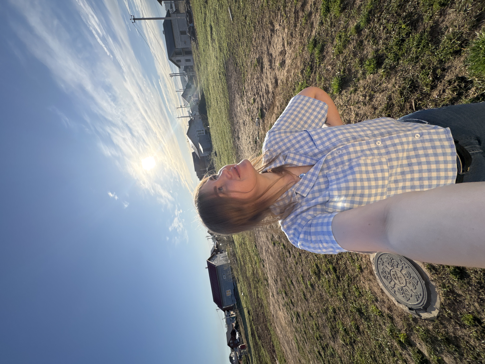 A young woman is in Ферма, Russia. The photo appears to be a selfie taken outdoors during the day in a rural or suburban area. The woman is wearing a blue and white checkered shirt and jeans. The setting appears to be a field or grassy area with houses visible in the background. A manhole cover is visible near her. The sky is blue with scattered clouds and the sun is visible in the distance.