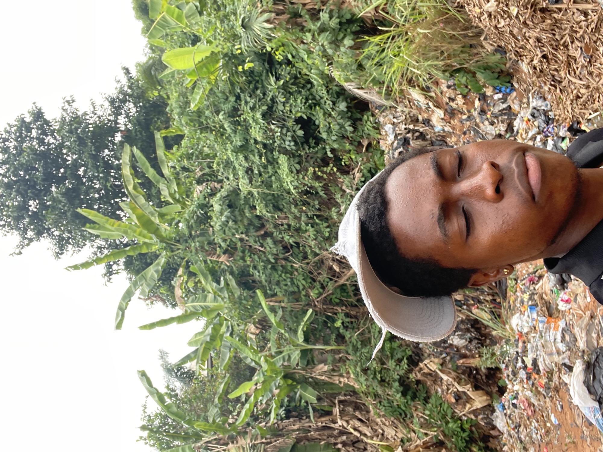 The image depicts a man with dark skin, wearing a light-colored cap and a dark shirt, eyes closed, seemingly lying down. He appears to be located in an area littered with trash and debris. In the background, lush green foliage, including banana trees, indicates a natural or semi-natural environment. The scene is suggested to be in Onitsha, Nigeria, based on the high amount of trash, common in areas of the city. The man's position and closed eyes suggest he might be sleeping or resting.