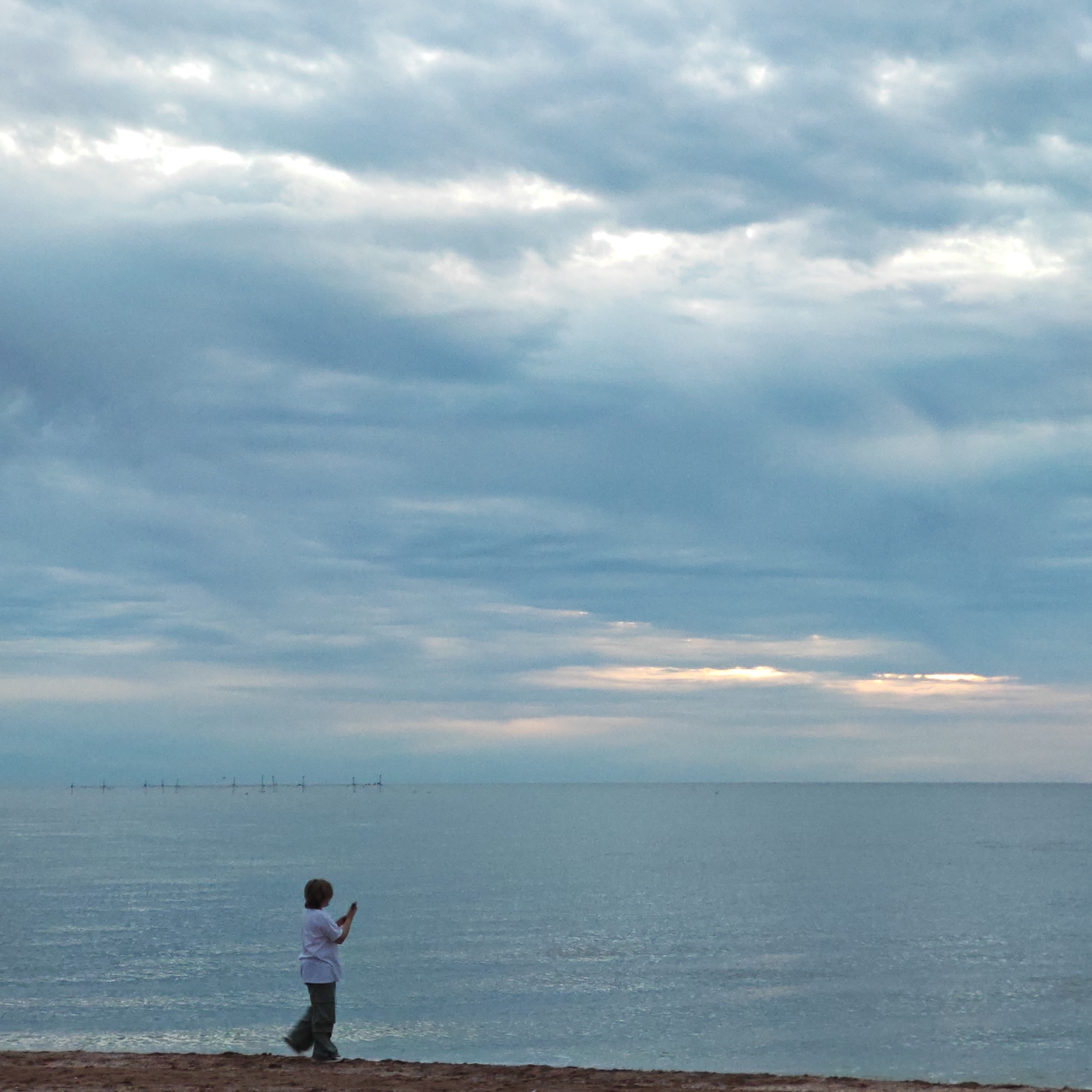 The image displays a coastal landscape featuring a lone individual, a large body of water, and an expansive sky. The scene is likely set at dawn or dusk, characterized by soft, diffused lighting and an overcast sky with patches of brighter light visible through the clouds near the horizon.
In the foreground, a sandy or earthy bank runs along the bottom, upon which a person, appearing to be a child or young individual, stands slightly to the right. Dressed in a light-colored short-sleeved top and dark trousers, they are holding a dark smartphone or similar electronic device with both hands, seemingly engrossed in its screen or using it to capture something.
The middle ground is dominated by a calm, blue-grey body of water, stretching towards the distant horizon. Along the horizon line, a series of small, dark, vertical structures, consistent with offshore wind turbines or other industrial installations, are faintly visible.
The sky occupies the majority of the frame, filled with layers of grey and blue-grey clouds. The overall mood of the image is quiet and contemplative. No visible text is present in the media. This location is identified as городское поселение Щелкино, Ukraine.