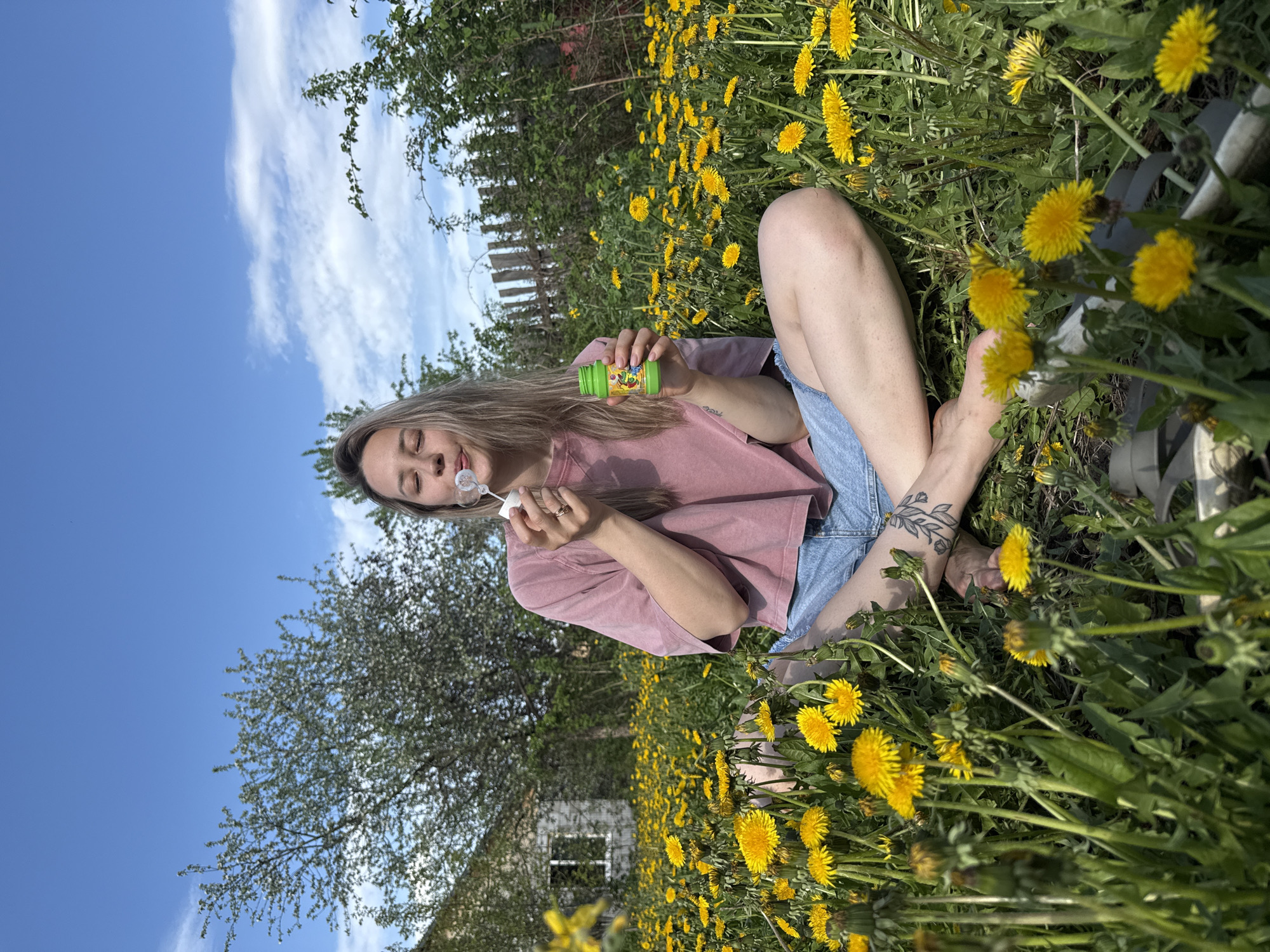 A woman is sitting in a field of dandelions, blowing bubbles. She is wearing a pink t-shirt and blue jean shorts, and she has a tattoo on her leg. The sky is blue with white clouds. There is a house and trees in the background. The location may be Chaikovsky, Russia.