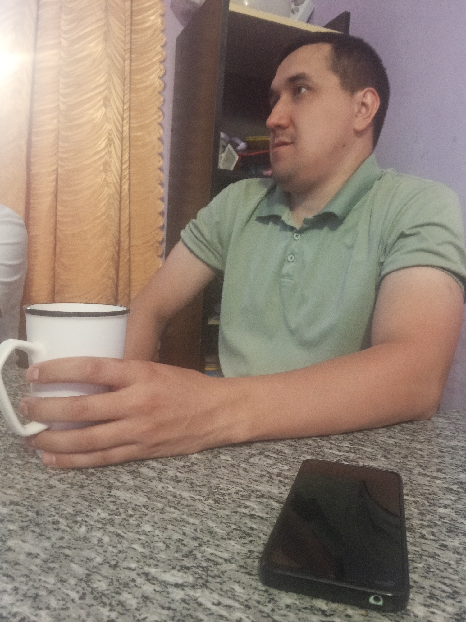 The image depicts an indoor scene, likely a room in a residential building in Chaikovsky, Russia. The primary subject is a man, seen from the waist up and slightly from the side, holding a white mug with a black rim. He is wearing a light green polo shirt with two buttons at the collar. His right arm is resting on a speckled granite-like surface, possibly a table. In the foreground, to the right of the mug, a black smartphone lies flat on the same surface. Behind the man, a shelf unit is visible, with some items on it. To the left, a golden, textured curtain hangs. The lighting suggests it is daytime, with natural light coming from the left, casting a slight glow on the curtain. The man appears to be looking off to the left of the frame.