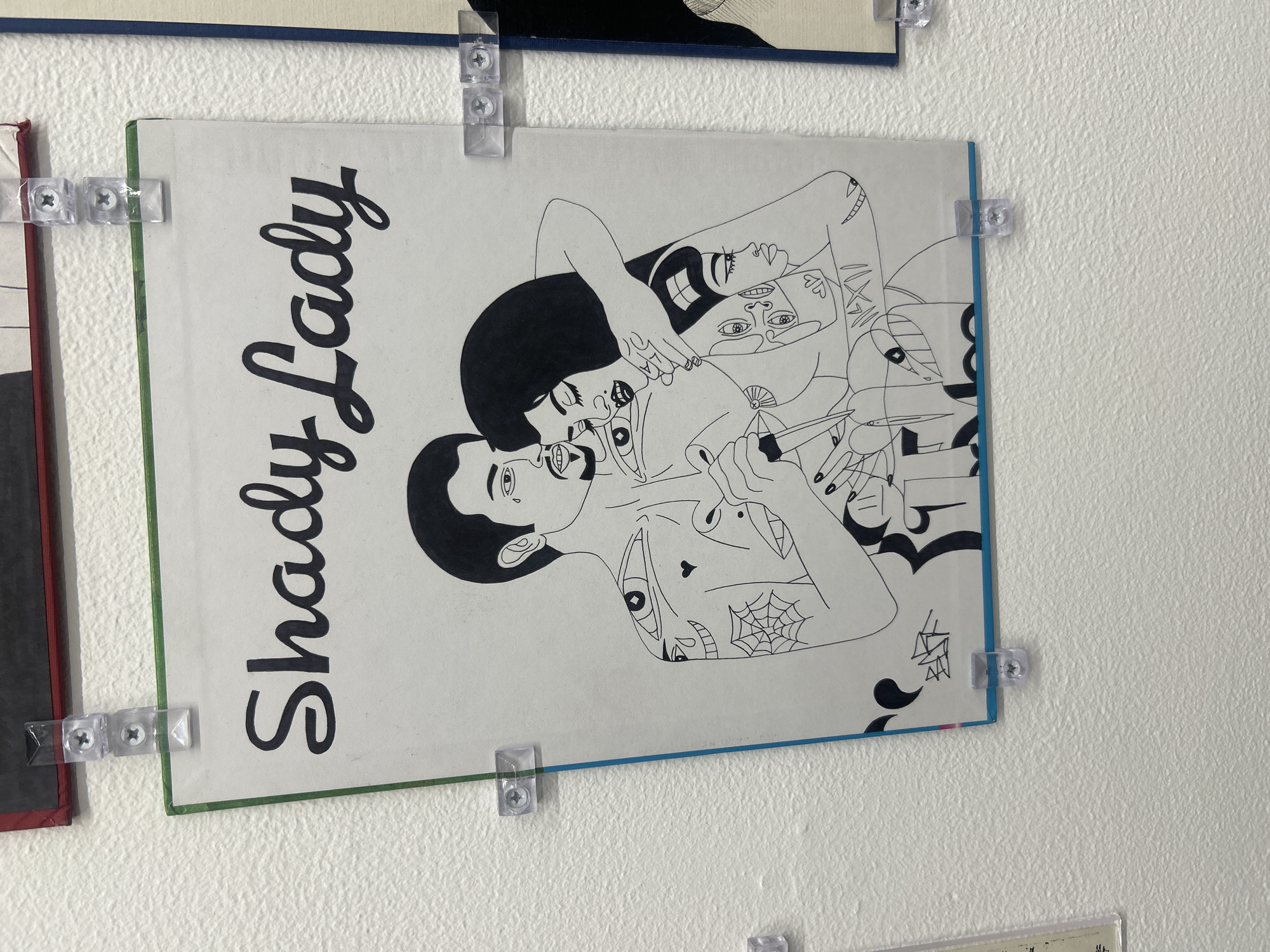 A framed artwork titled "Shady Lady" hangs on a white wall, secured with clear plastic clips and screws. The drawing depicts two figures in black and white, with tattoos and stylized features, reminiscent of an illustration. The artwork's frame is green on the left and blue on the right. The wall is lightly textured. The location is in Los Angeles, United States of America, inside an art gallery.