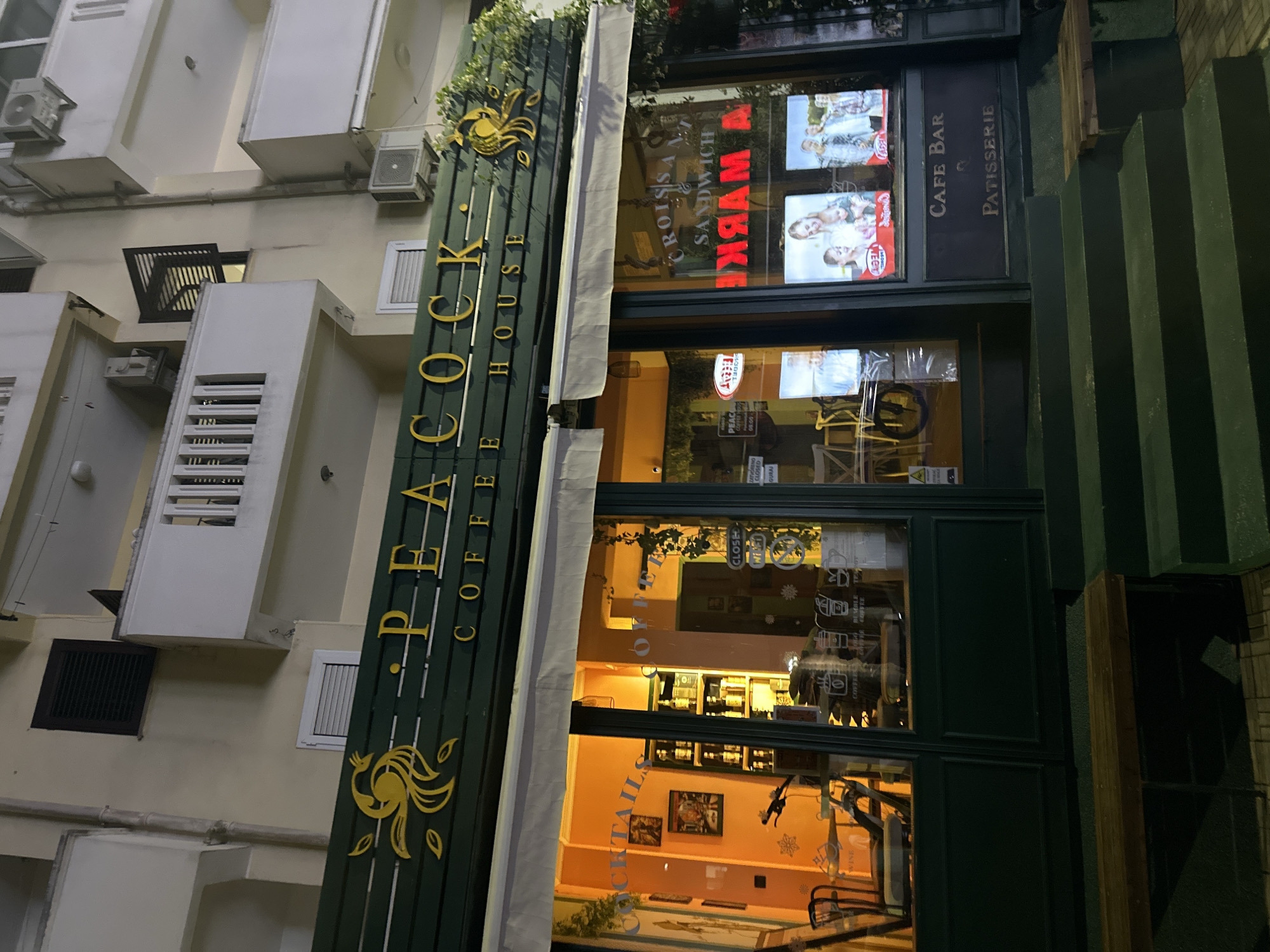 The image shows the storefront of "Peacock Coffee House," a café bar and patisserie. The sign is a long, green panel with the name in gold lettering and a decorative gold peacock design. The building facade is light-colored and has air conditioning units and windows. The cafe's interior is visible through the windows, with warm lighting and displays of drinks and goods. The reflected words 'A MARK' with a sandwich are visible through the windows. The location is suggested to be in the city of Budva, Montenegro.