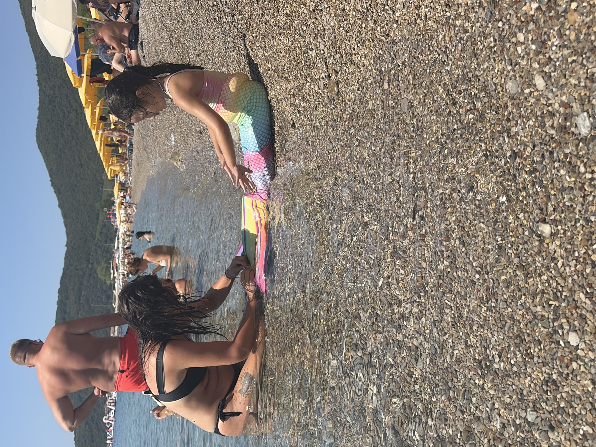 The image depicts a sunny day at a pebble beach in Prijevor, Montenegro. In the foreground, a woman in a black bikini is seated in the shallow water, holding hands with a young girl wearing a rainbow-colored mermaid tail costume. The girl is leaning forward, her hands reaching towards the water. Behind them, a man with blonde hair and red shorts stands in the water, facing away from the camera. The background shows a busy beach scene with numerous yellow and blue umbrellas, people sunbathing, and the clear blue sea stretching towards a tree-covered hillside. The water is clear and shallow, revealing the pebble-covered seabed. The sun appears to be high in the sky, casting shadows and indicating midday. The overall atmosphere is one of leisure and enjoyment on a summer vacation.