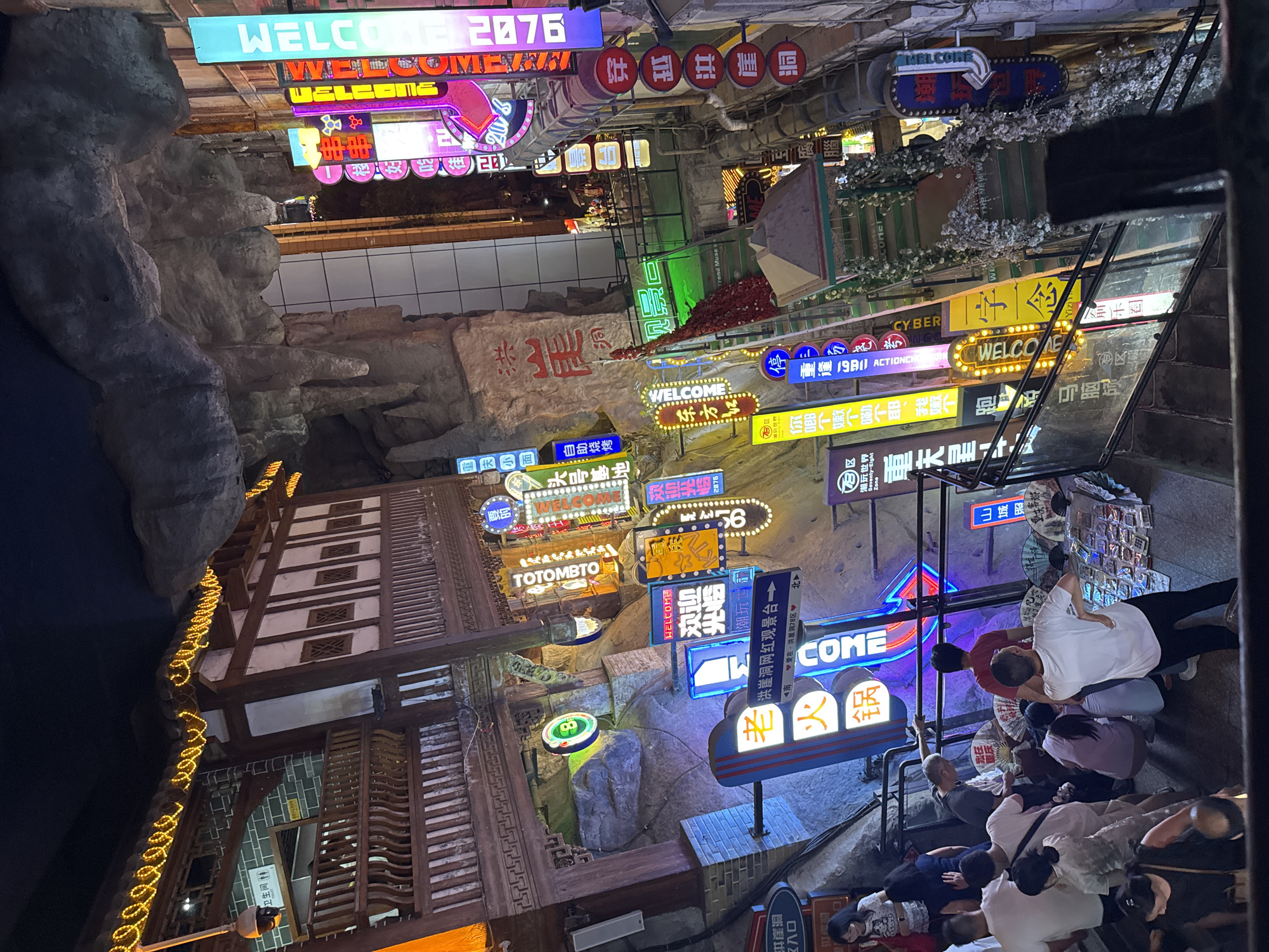The image depicts a bustling night scene in a multi-level indoor marketplace or entertainment complex, likely located in Yuzhong District, China. The architecture features traditional Chinese elements with wooden structures, stone facades, and a rock-like ceiling, creating a cave-like or historical atmosphere. Numerous neon signs in Chinese and English, including "WELCOME," illuminate the space, advertising shops, restaurants, and attractions. People are seen navigating through the crowded walkways and staircases, some looking at merchandise displayed on tables, while others are in conversation. The overall impression is one of vibrant commercial activity and a lively social gathering.