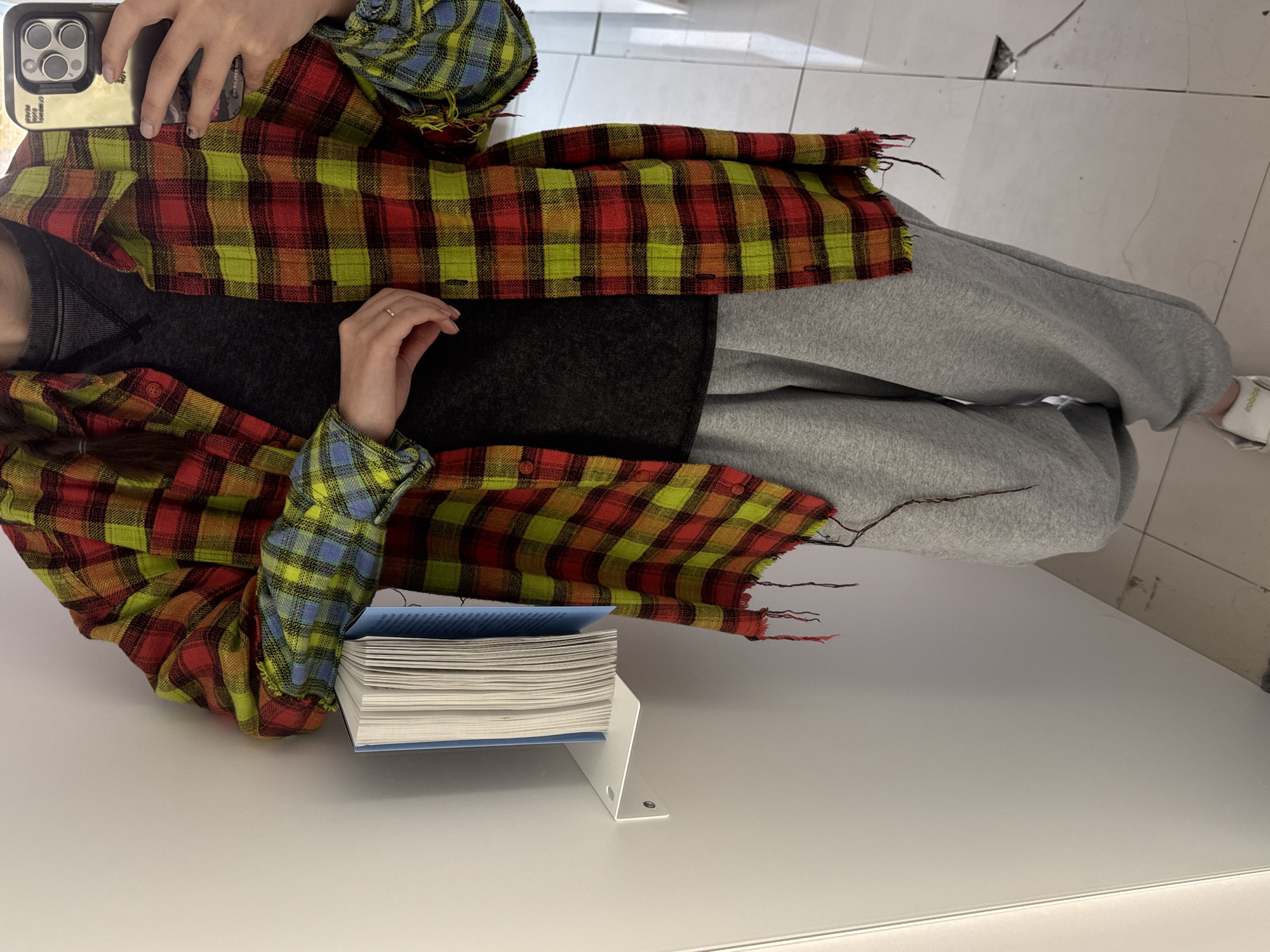 A person sits at a white desk in an indoor setting, holding a mobile phone and wearing a plaid shirt over a dark-colored top and gray sweatpants. A book rests on a white bookstand. The location is in the city of Unknown, Unknown.