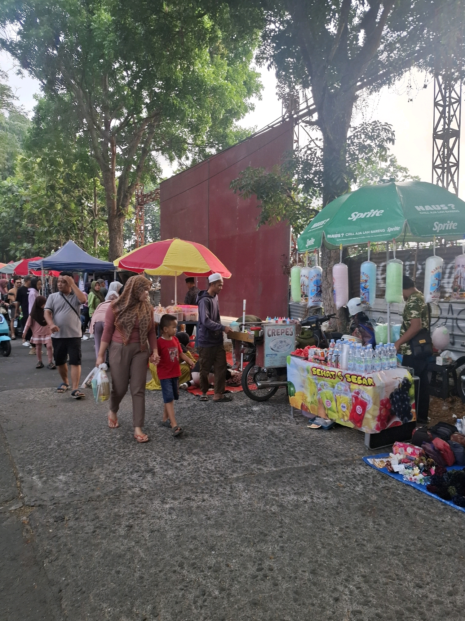 The image depicts a bustling street scene in Karangtengah, Indonesia, likely a local market or fair. In the foreground, a woman wearing a patterned headscarf and a man in a gray t-shirt and shorts are walking, the woman holding a plastic bag of groceries and the man adjusting his sunglasses. A young boy in a red t-shirt and blue shorts walks hand-in-hand with the woman. To the right, a street vendor is operating a stand under a green Sprite umbrella, selling drinks and possibly other refreshments, with various bottles of water displayed on a table covered with a colorful cloth depicting fruit. Behind this stand, other vendors are set up, including one with a "Crepes" sign on a cart. A red wall forms a backdrop for some of the stalls, and large trees with lush green foliage line the street. In the background, more people can be seen browsing or walking through the market. The overall impression is one of lively community commerce taking place outdoors.