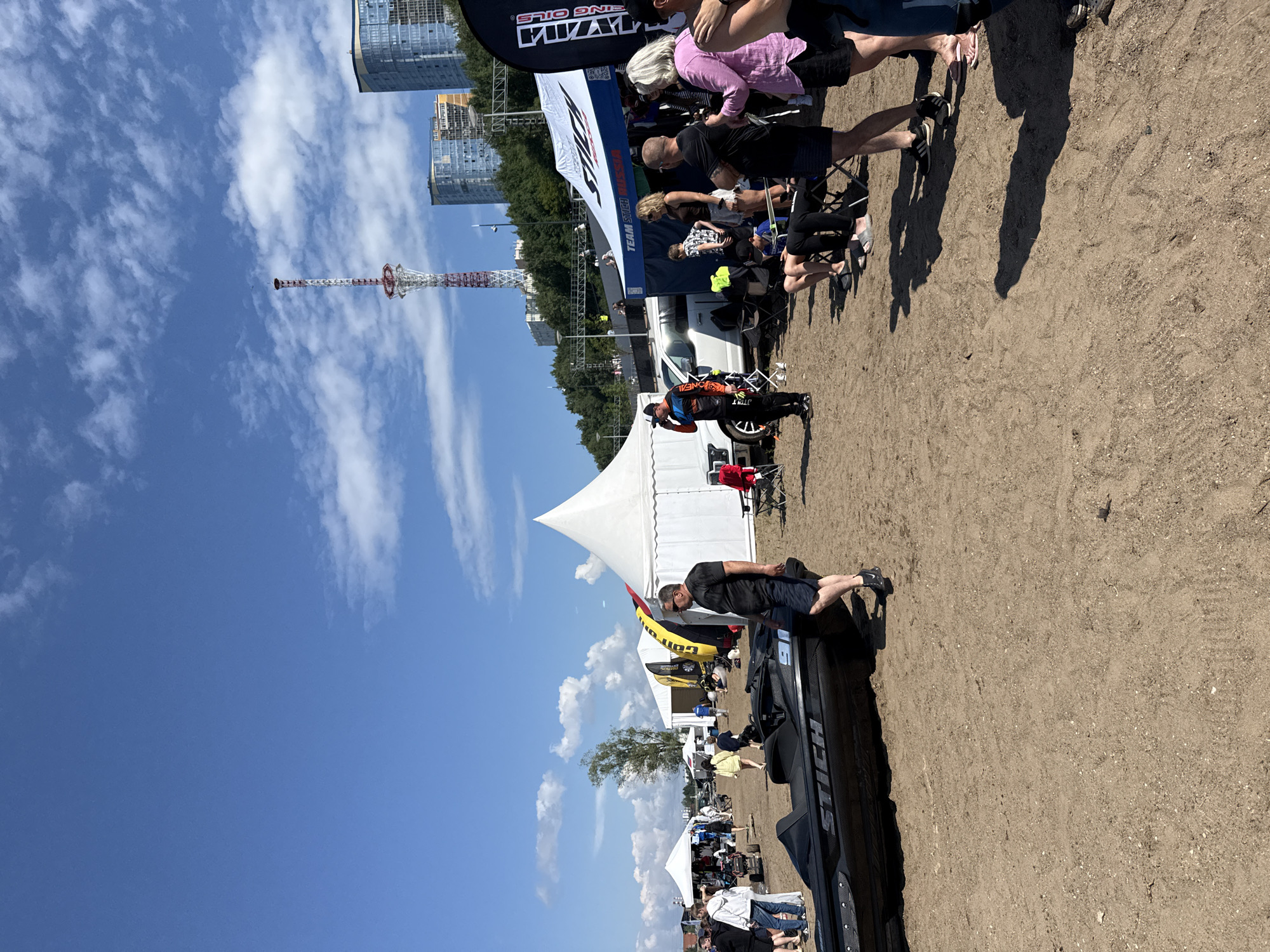 An outdoor event is taking place on a sandy beach or area, with several tents set up and a large crowd of people milling around. In the foreground, a black jet ski with the word "STICH" printed on its side is positioned on the sand. Several people are standing near the jet ski and in the background, observing the event. Among the tents, one appears to be a medical tent, with "TEAM STICH RUSSIA" visible on another. A white pickup truck is parked near one of the tents. In the distance, tall buildings and a radio tower are visible against a blue sky with scattered clouds. The scene suggests a recreational or sporting event. The location is the city of Perm, Russia, along the Kama River.