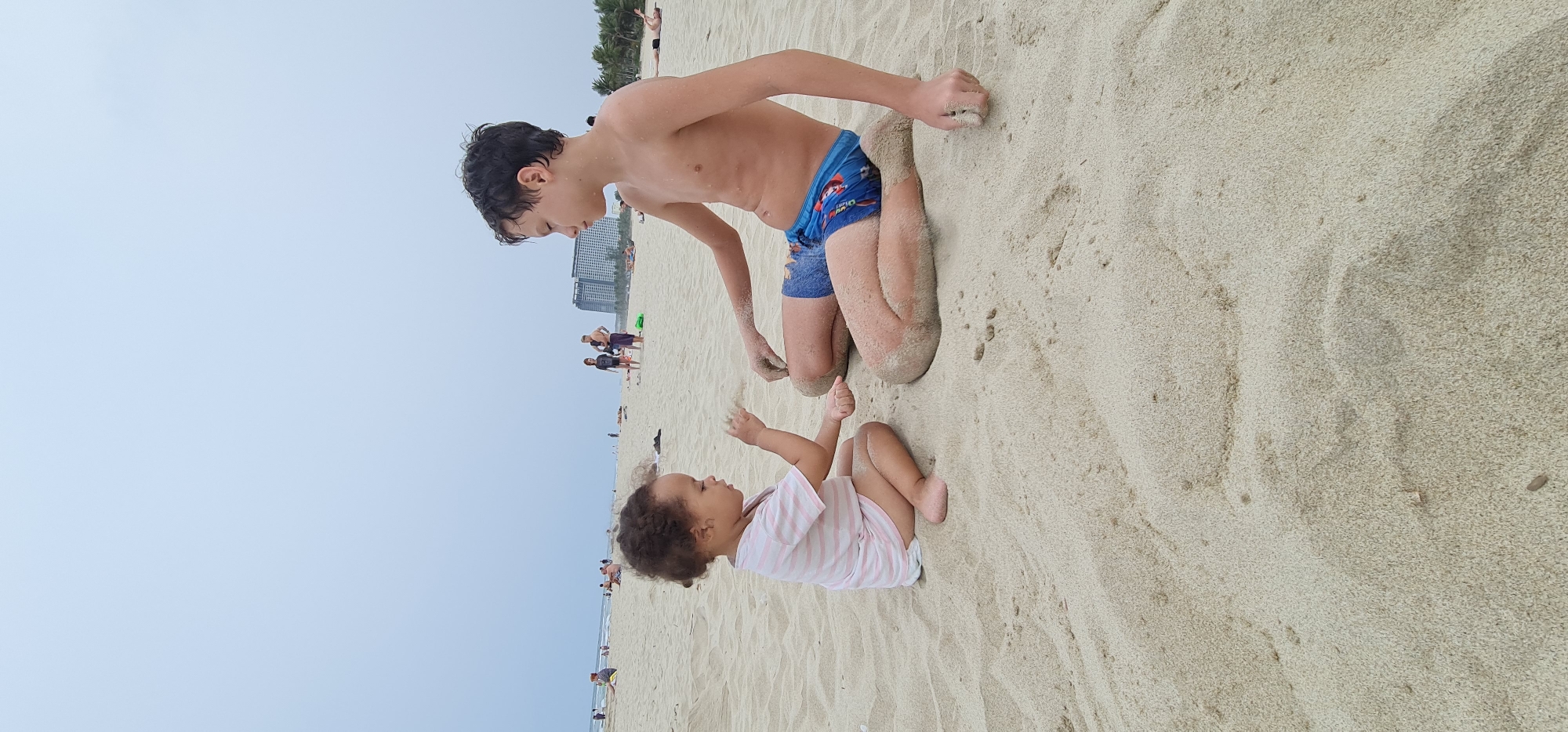 The image depicts a young boy and a toddler playing on a sandy beach in Đà Nẵng, Vietnam. The older boy, shirtless and wearing blue swim trunks, kneels in the sand, with sand covering his legs. The younger child, wearing a pink and white striped shirt, sits facing him, with her arms raised. The beach is populated with other people in the background, including some figures closer to the water and others near buildings. The sky is overcast. The foreground shows undisturbed sand.