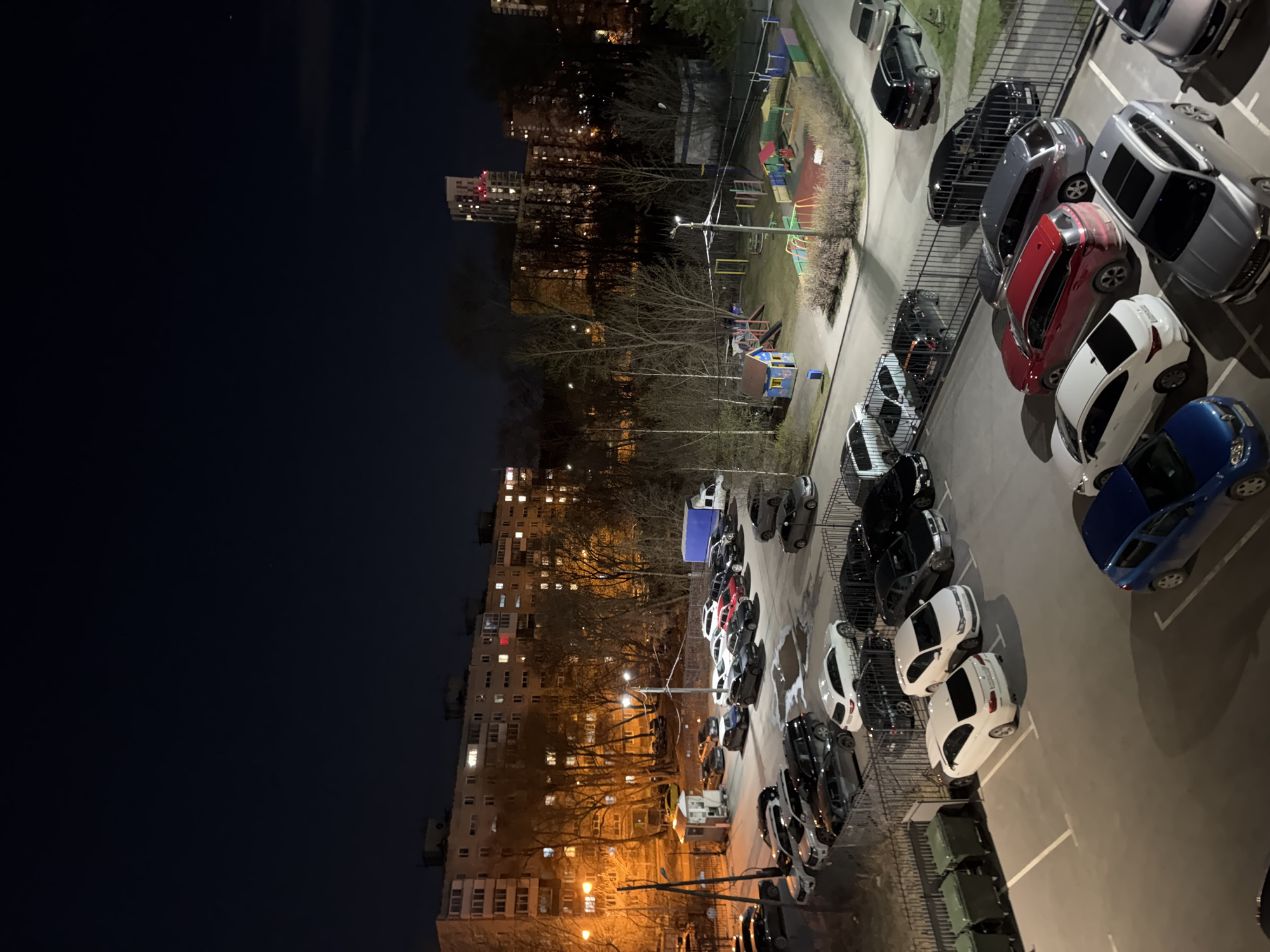 This media file depicts an aerial view of a residential area at night, likely captured from a high vantage point such as an upper-story apartment window. The scene is dominated by numerous parked cars, multi-story buildings, and a playground, all illuminated by artificial lights against a dark night sky.
In the foreground and extending into the mid-ground, a large parking lot is visible, filled with a diverse array of cars, primarily sedans and SUVs, in colors such as white, red, blue, grey, and black. Many vehicles are neatly parked within marked spaces. To the left of the parking area, a street or access road is visible, with more cars parked alongside. Metal fencing separates different sections of the parking and residential areas. A row of green garbage bins is seen in the bottom right corner.
Further into the mid-ground, a colorful children's playground with slides, swings, and climbing frames is present, also enclosed by fencing. Several bare trees, suggesting late autumn or winter, are scattered throughout the green spaces separating the parking and building areas. Beyond the playground, multi-story residential buildings line the horizon, with many windows lit, indicating occupancy. One prominent building in the distance appears to be under construction or features a tall structure with red lights on top. Another large apartment complex with numerous illuminated windows occupies the entire left side of the frame, towering over the scene.
The entire area is lit by streetlights, casting distinct shadows and reflections, particularly on the parked cars. The dark sky confirms it is nighttime. There is no visible activity, with the scene appearing static. No discernible text is visible on any signs, vehicles, or buildings. The architecture and urban layout are consistent with residential areas found in cities like Perm, Russia.