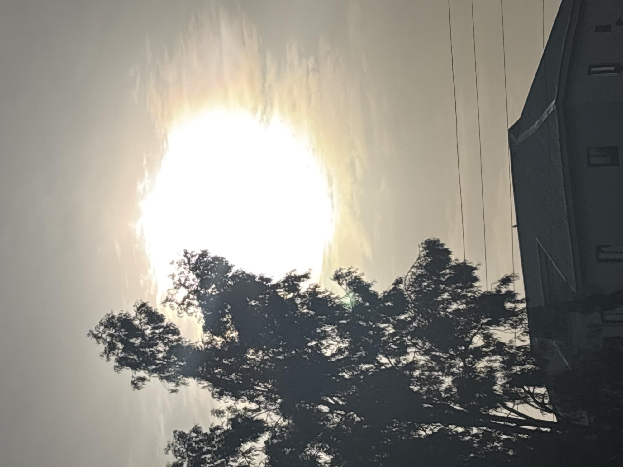 The image depicts a silhouetted tree in the foreground, its branches extending upwards and to the left against a hazy sky. The sun is a bright, luminous orb in the upper right quadrant, partially obscured by clouds that create a soft halo effect. In the lower half of the image, the roofline of a building is visible, with a dark, metallic surface. A few power lines stretch horizontally across the sky above the building. The overall scene is consistent with an outdoor environment in Bukuru, Nigeria, possibly during sunrise or sunset, given the diffused light and the atmospheric haze. There are no people or specific actions clearly identifiable in the image.