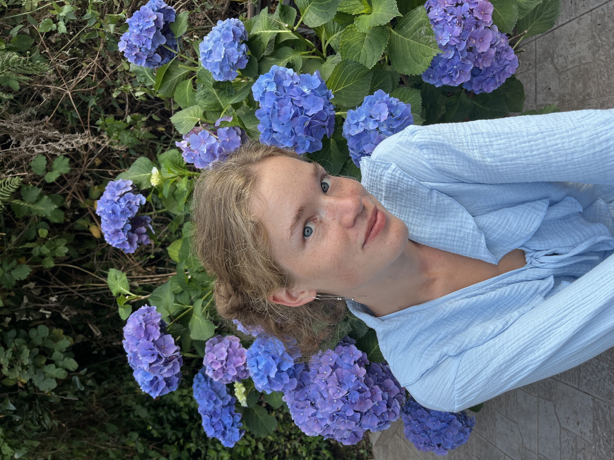 In Budva, Montenegro, a young woman with fair skin, freckles, and light brown hair pulled up in a bun is photographed outdoors. She is wearing a light blue, crinkled fabric shirt with a ruffled neckline that ties in the front. She has a silver earring in her visible left ear. She is positioned in front of a large hydrangea bush with abundant blue and purple blooms. The background consists of greenery and tangled branches. The ground beneath her appears to be paved with stone tiles. The woman is looking directly at the camera with a slight smile.