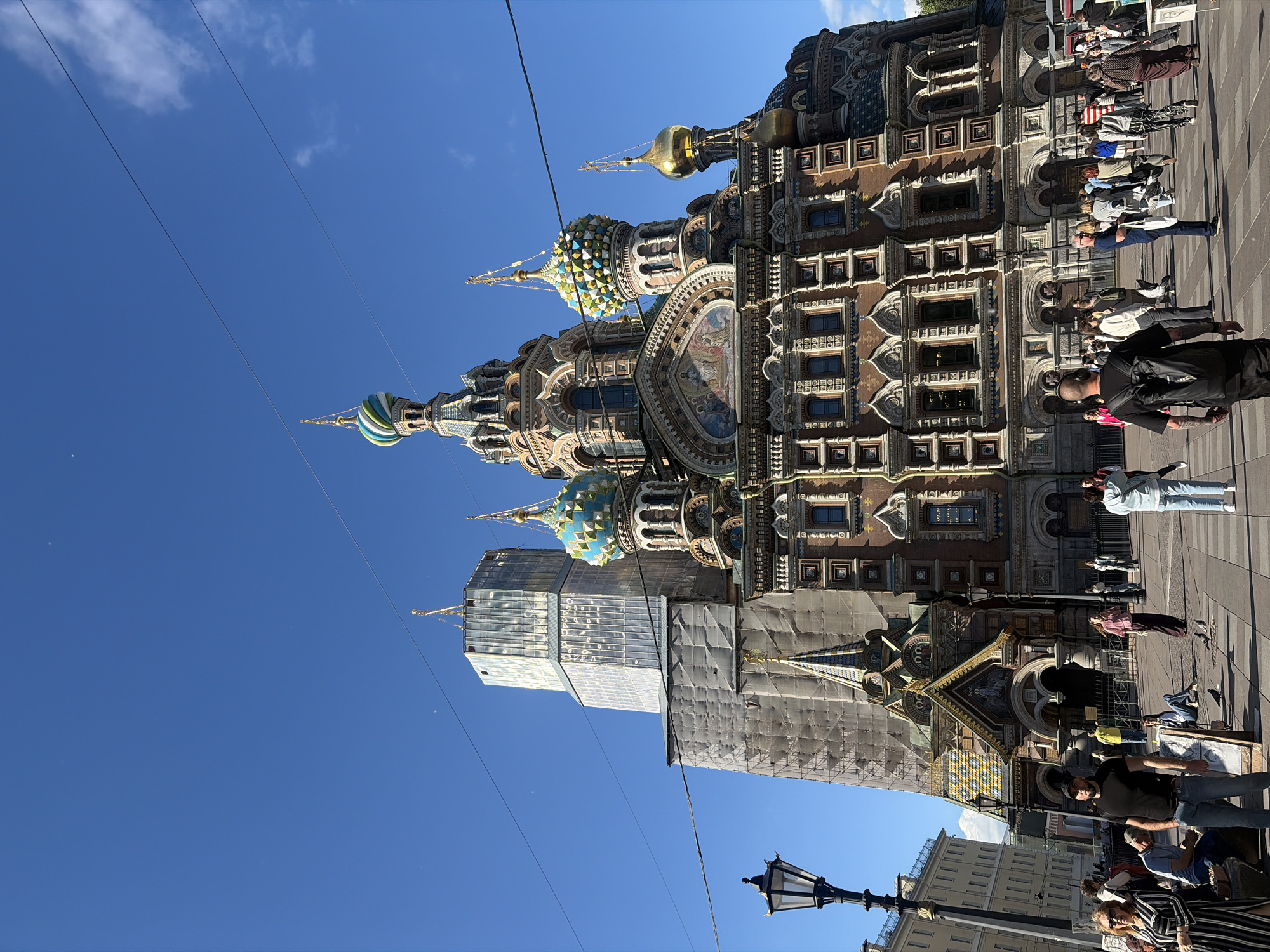 The image shows a view of the Church of the Savior on Spilled Blood in Saint Petersburg, Russia, on a clear, sunny day. The iconic church, with its colorful onion domes and intricate facade, is partially obscured by scaffolding and a large, modern structure on its left, suggesting ongoing renovation or construction. A bright blue sky with a few wispy clouds forms the backdrop.
In the foreground, a paved public square is filled with people. Some individuals are walking, while others are sitting or standing. A man in a black t-shirt and jeans is prominently positioned in the mid-ground, facing towards the camera. To his left, a woman in a striped dress is also visible. Several pigeons are walking on the pavement. A traditional black lamppost stands to the left of the church entrance. The surrounding buildings in the background appear to be of classical architecture. Power lines stretch across the upper portion of the image.