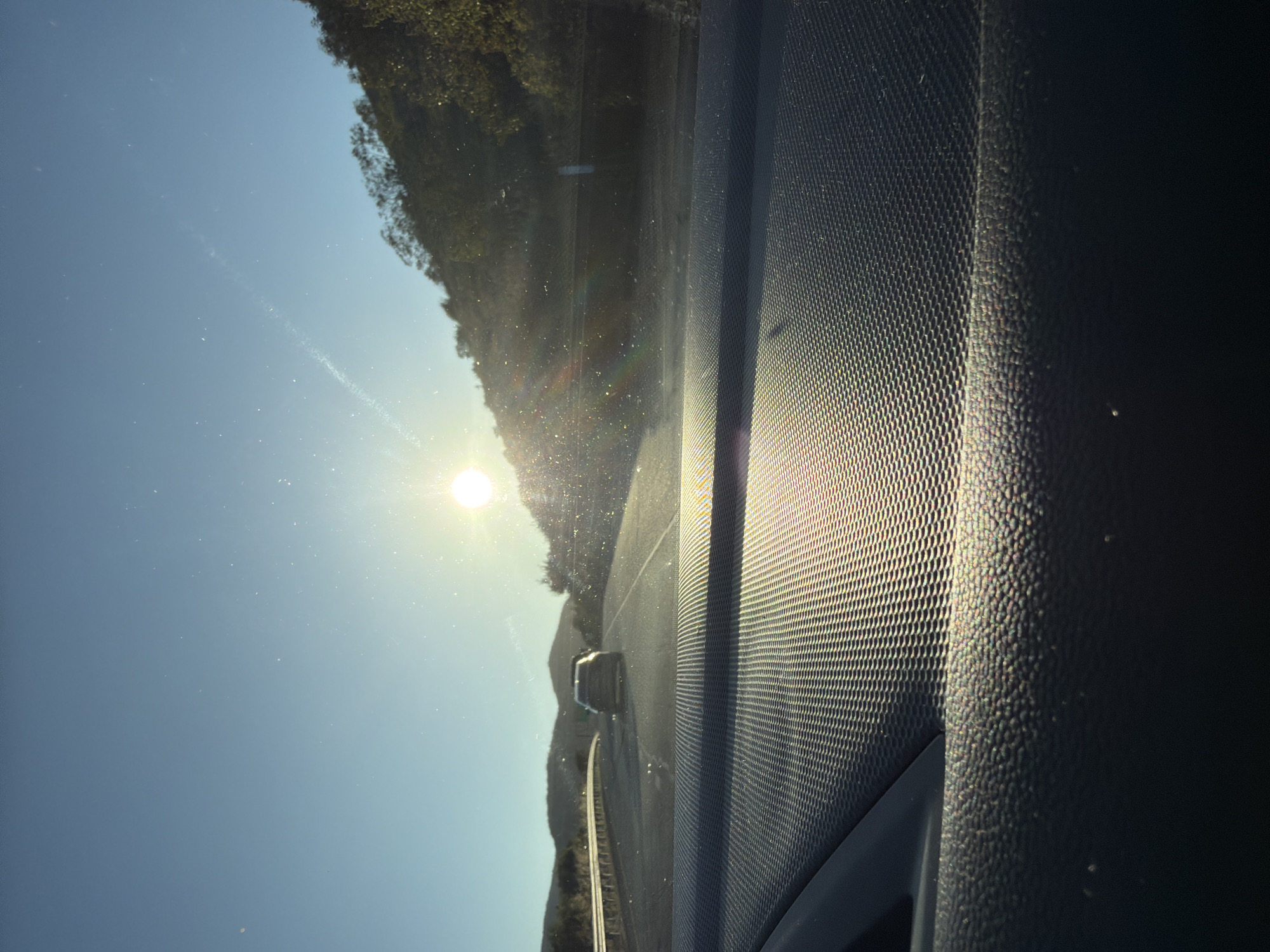 The image captures a view from inside a vehicle traveling on a highway, likely a coastal road, in Laguna Beach, California, United States of America. The sun is low in the sky, casting a bright glare and creating lens flares across the scene. The road ahead is visible, with a single car in front. On either side of the highway, there are hills covered with vegetation. The dashboard of the vehicle is in the foreground, with its textured surface reflecting the sunlight. The sky is a clear blue, with a few faint streaks suggesting contrails or atmospheric haze. The overall impression is one of a scenic drive during golden hour.