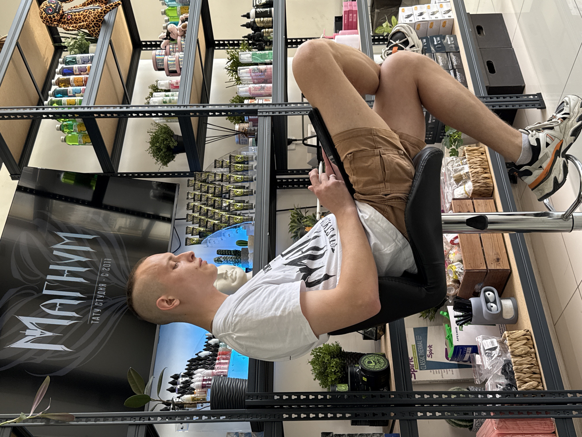 The image shows a person sitting backwards on a black chair, with their legs crossed and feet resting on the chair's footrest. They are holding a black tablet in their hands. The person is wearing a white t-shirt and brown shorts. They have short, light-colored hair. Behind the person is a shelving unit filled with various items, including bottles, plants, and boxes. A poster with the text "ΜΑΓΗΥΜ TATY CTУДИЯ - С 2011", which translates to "MAGNUM TATTOO STUDIO - C 2011", is partially visible. The presence of these items and the text on the poster suggest that the location might be a tattoo studio, likely in Rostov-on-Don, Russia, given the Cyrillic alphabet used in the poster text.