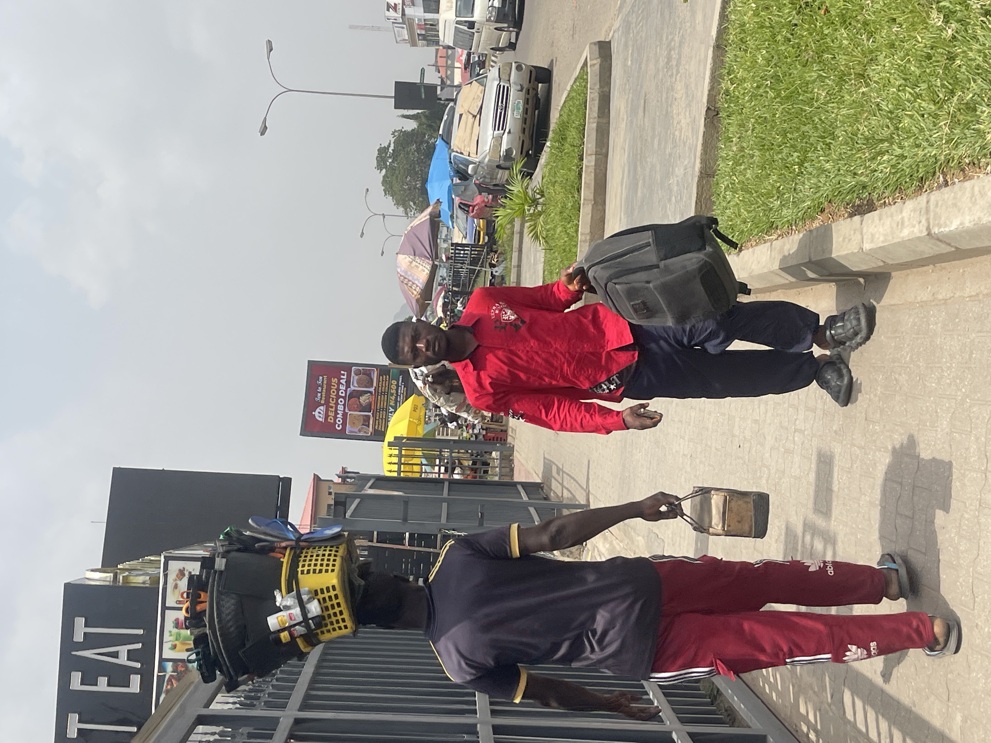 This photograph captures a vibrant street scene in Abuja, Nigeria, during bright daylight hours. The sky is largely clear with some wispy clouds. The image is taken at a slight upward angle, showing a busy urban environment.
In the foreground, two men are walking. The man on the left is seen from behind, carrying a yellow plastic basket filled with various items, including bottles and what appears to be flip-flops, balanced on his head. He wears a dark blue short-sleeved shirt with yellow trim and maroon Adidas track pants with white stripes and logos, along with flip-flops. He holds a small, light brown rectangular bag in his right hand. To his right, another man, facing partially towards the viewer, carries a dark grey or black backpack over his left shoulder. He wears a red long-sleeved shirt with a crest-like logo on the chest and dark trousers, paired with dark sneakers.
The background reveals a bustling street lined with cars and vans, some appearing to be parked or stalled, with one silver SUV having cardboard covering parts of its roof or windscreen. Colorful market umbrellas (blue, yellow, and patterned) are visible, indicating the presence of market stalls or street vendors further down the road. Streetlights with multiple arms extend into the sky.
On the far right, a concrete sidewalk borders a well-maintained grassy median. On the far left, a dark-colored building features large white lettering that reads "TEAT" (likely part of a larger word like "Restaurant" or "EAT"). Higher up and further back, a prominent billboard advertises "Tea To Fun Restaurant" with text "DELICIOUS COMBO DEAL!" and "ONLY N4,500," alongside images of food. The overall scene depicts a typical active day in a Nigerian city, with people engaged in commerce and daily movement.