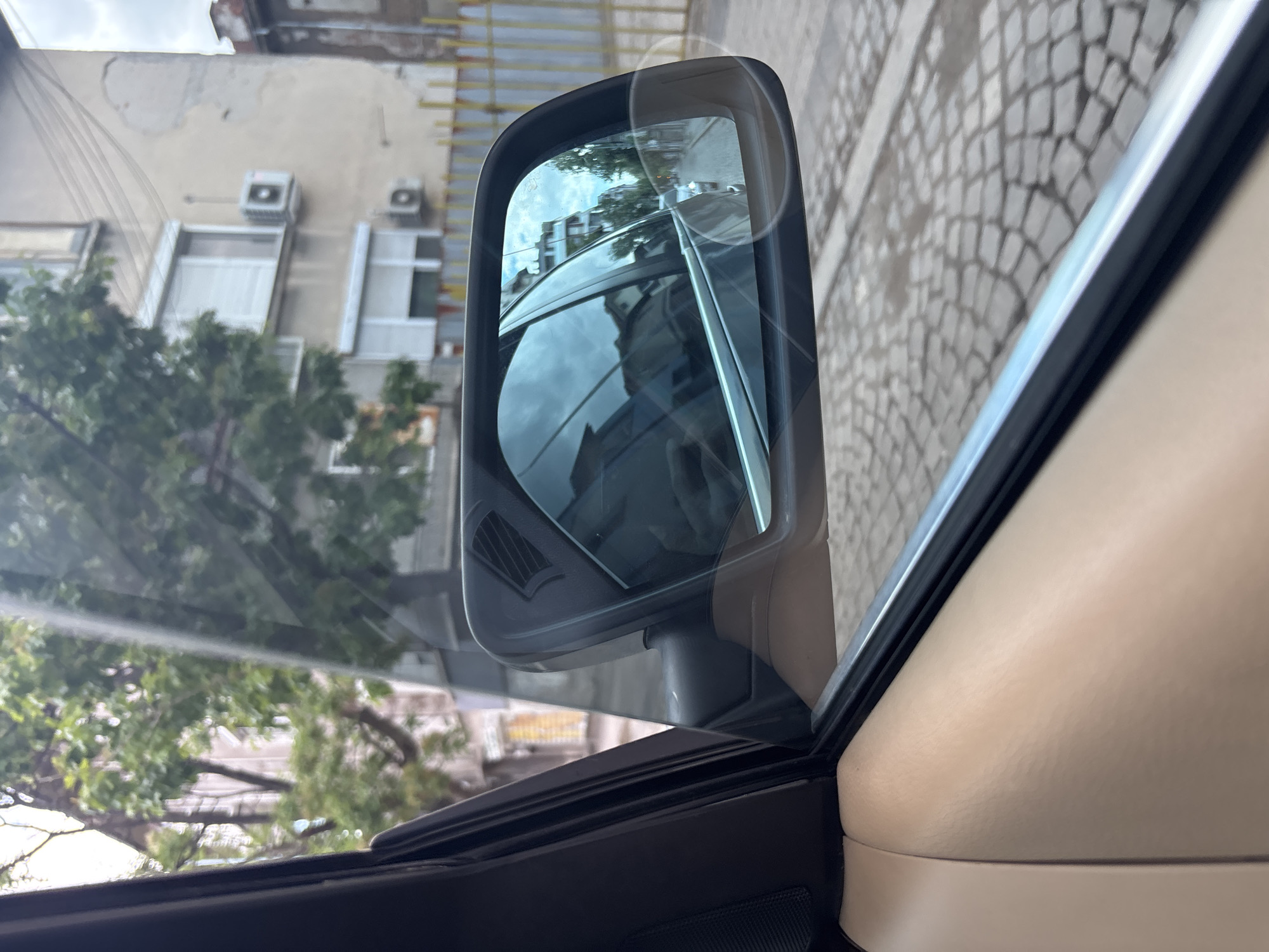 The image shows a side mirror of a car, reflecting a street scene in Plovdiv, Bulgaria. The reflection reveals a building with multiple windows and air conditioning units, along with trees and a patterned cobblestone road. The car's interior is visible, including parts of the dashboard and window frame in light beige and black colors. The viewpoint is from inside the car, looking towards the side mirror.