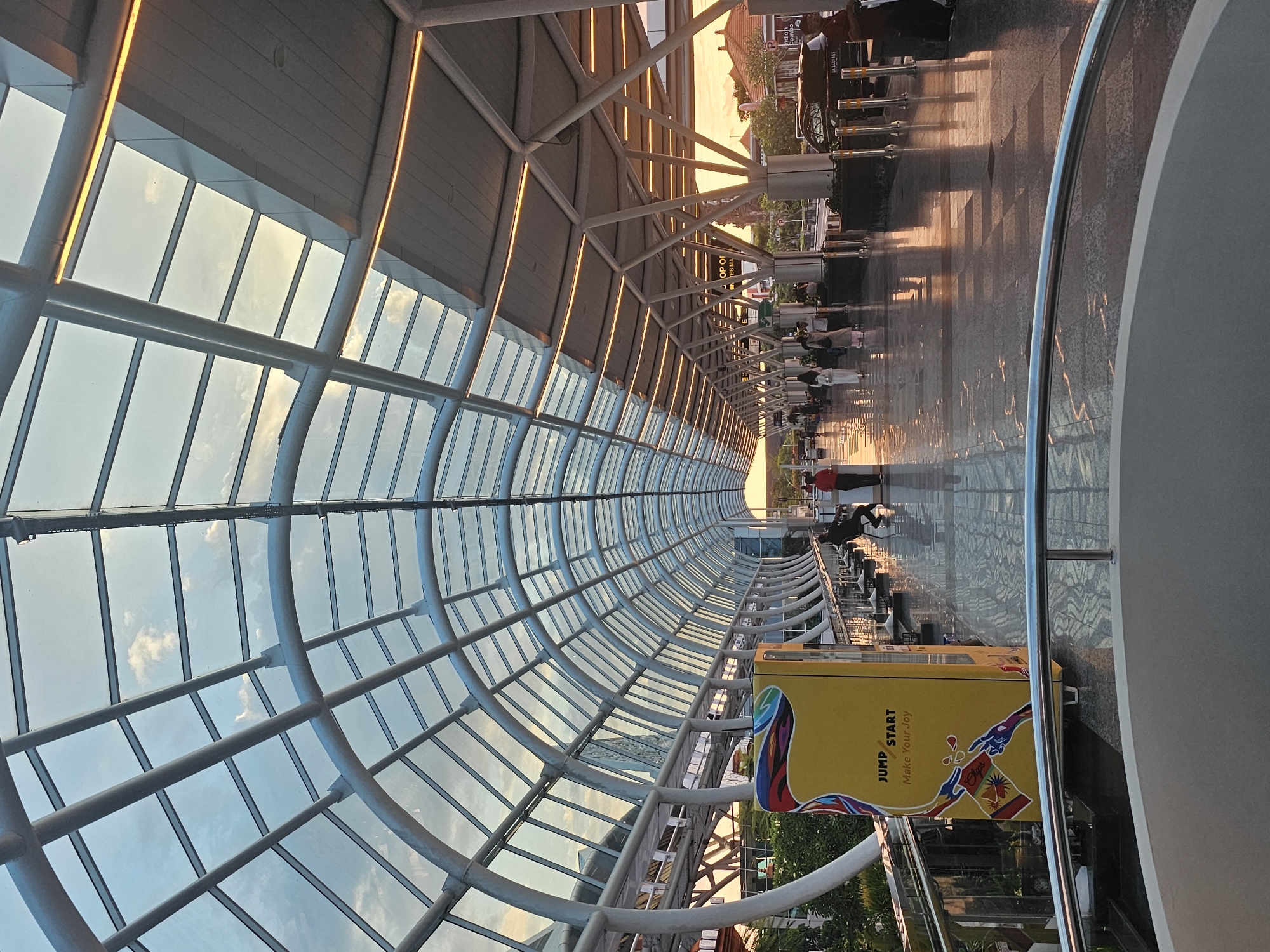 The image shows the interior of a covered walkway or bridge, likely at an airport. The structure has a curved roof made of glass panels supported by a steel framework. Outside the glass roof, the sky is visible. The walkway floor is patterned and reflects light, creating a sheen. People are walking along the walkway, and some are seated at tables. A yellow advertisement kiosk is visible on the right side of the walkway. The image shows the Domestic Terminal at I Gusti Ngurah Rai International Airport in Kuta, Indonesia.