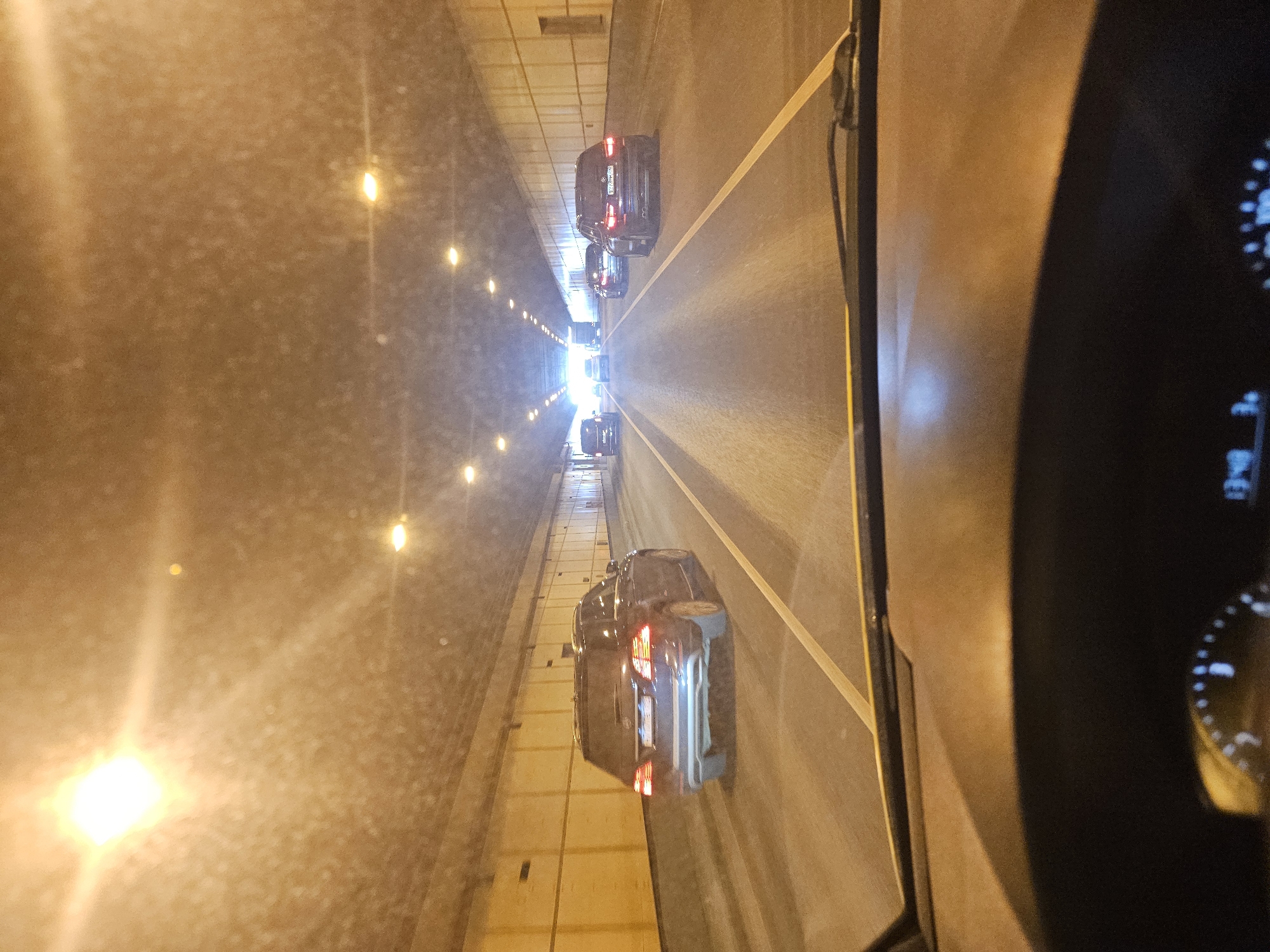 The image depicts a view from inside a vehicle driving through a tunnel, likely in the city of Khimki, Russia, based on the license plate of a car in front. Several cars are visible ahead, all traveling in the same direction. The tunnel walls are lined with lights, and a yellow line marks the lane. The dashboard of the vehicle taking the picture is partially visible on the right side of the frame.