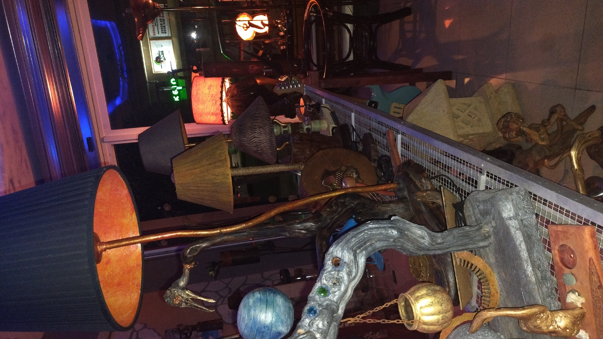 The image depicts a cluttered display of artistic objects, likely in a shop or gallery. The scene is dimly lit with warm and cool lighting, suggesting it might be evening or an interior space with artificial illumination. The overall impression is one of eclectic and possibly handmade items.
Prominently featured are several lamps with varying lampshades, including a large dark blue pleated shade with an orange interior and a conical gold lampshade. Interspersed among the lamps are various sculptural elements made of wood, metal, and possibly stone. One striking sculpture appears to be a twisted, weathered tree branch adorned with colorful glass or gem-like accents. Other metallic sculptures, some gold-toned and others darker, are also visible. A golden pot hanging from a chain adds to the decorative arrangement. In the background, a neon sign that appears to read "KEIO" in green letters is visible through a window, along with some indistinct signage and the reflection of blue lights. The presence of a mesh barrier suggests a display area or a separation between viewing and display. The style of the items and the overall ambiance suggest a creative, perhaps bohemian, or artistic setting. There are no clear indications of people or a specific event taking place, but rather a static display of objects.