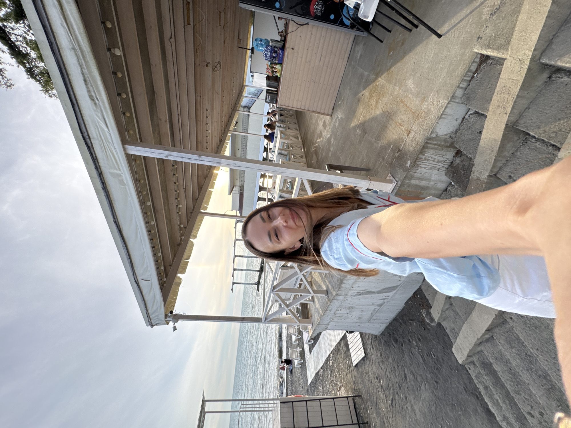 A woman takes a selfie on a beach in Sochi, Russia. She is standing on concrete stairs leading down to a dark sandy beach. To her left, the Black Sea is visible with gentle waves. A white wooden fence separates the beach from a covered outdoor dining area to her right. Several tables and chairs are set up in the dining area, with a few patrons seated. The woman is wearing a light blue top with red trim and white pants. The sun is setting, casting a warm glow on the scene.