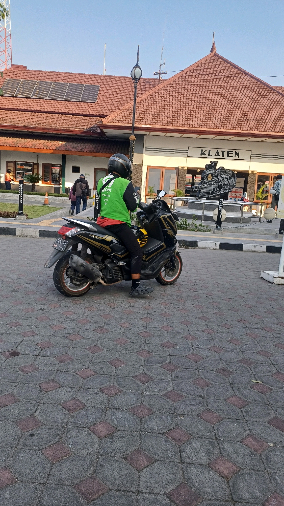 A person wearing a green jacket with "Gojek" branding and a black helmet is sitting on a black motorcycle in front of the Klaten train station in Klaten, Indonesia. The station building has a red tiled roof and a sign that reads "KLATEN." In front of the station is a decorative sculpture of a steam train. The ground is paved with patterned interlocking concrete blocks. The sky is clear and blue. There are other people and some vegetation visible in the background.