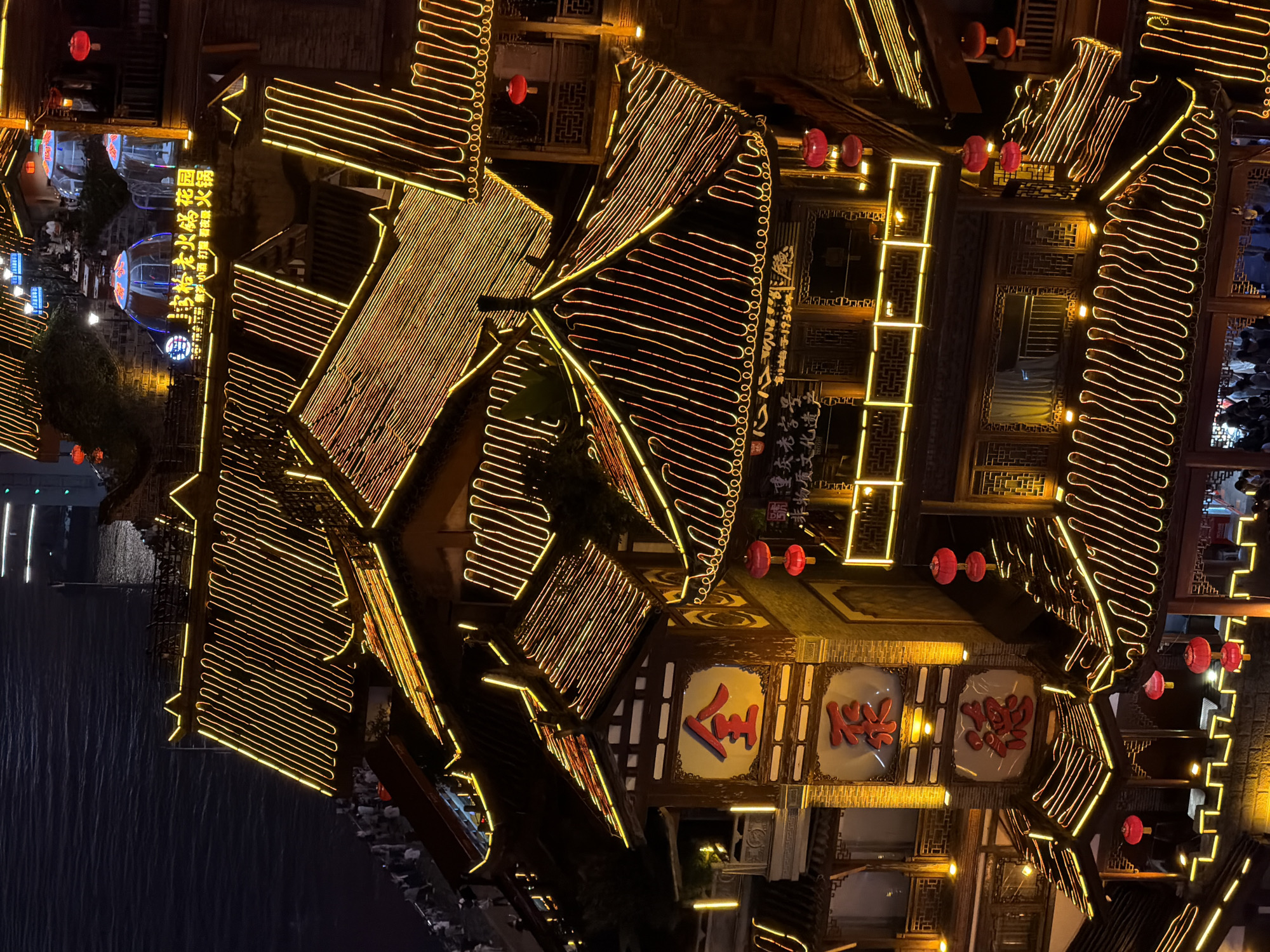 The image shows an aerial view of a busy night scene in Yuzhong District, Chongqing, China. The architecture features traditional Chinese-style buildings with multi-tiered, dark tiled roofs. These buildings are extensively decorated with bright yellow neon lights tracing the eaves and structural lines, creating a vibrant and festive atmosphere. Red lanterns are also strung throughout the scene, adding pops of color against the dark night.
In the foreground, a prominent building displays large red characters. The topmost character, in a circular frame, appears to be "金" (jīn), meaning gold. Below it, in a rectangular frame, is the character "泰" (tài), meaning peace or great. Further down, another large red character, "德" (dé), meaning virtue or morality, is visible. These characters suggest the name or theme of the establishment.
Below these prominent signs, more traditional wooden structures with intricate latticework windows are visible. The roofs themselves are illuminated, highlighting their layered construction. In the middle ground, a sign with Chinese characters and illuminated domes suggests a dining area, possibly for hot pot, as "火锅" (huǒguō - hot pot) is visible on the sign. Several small, clear dome-like structures are set up, likely for outdoor dining.
Numerous people are visible, walking along pathways and gathered in and around the buildings. They appear to be tourists or locals enjoying the evening. The buildings are situated close to a body of water, which is dark and reflects the distant city lights. The overall impression is one of a lively entertainment and dining district, illuminated by a multitude of lights against the night sky.