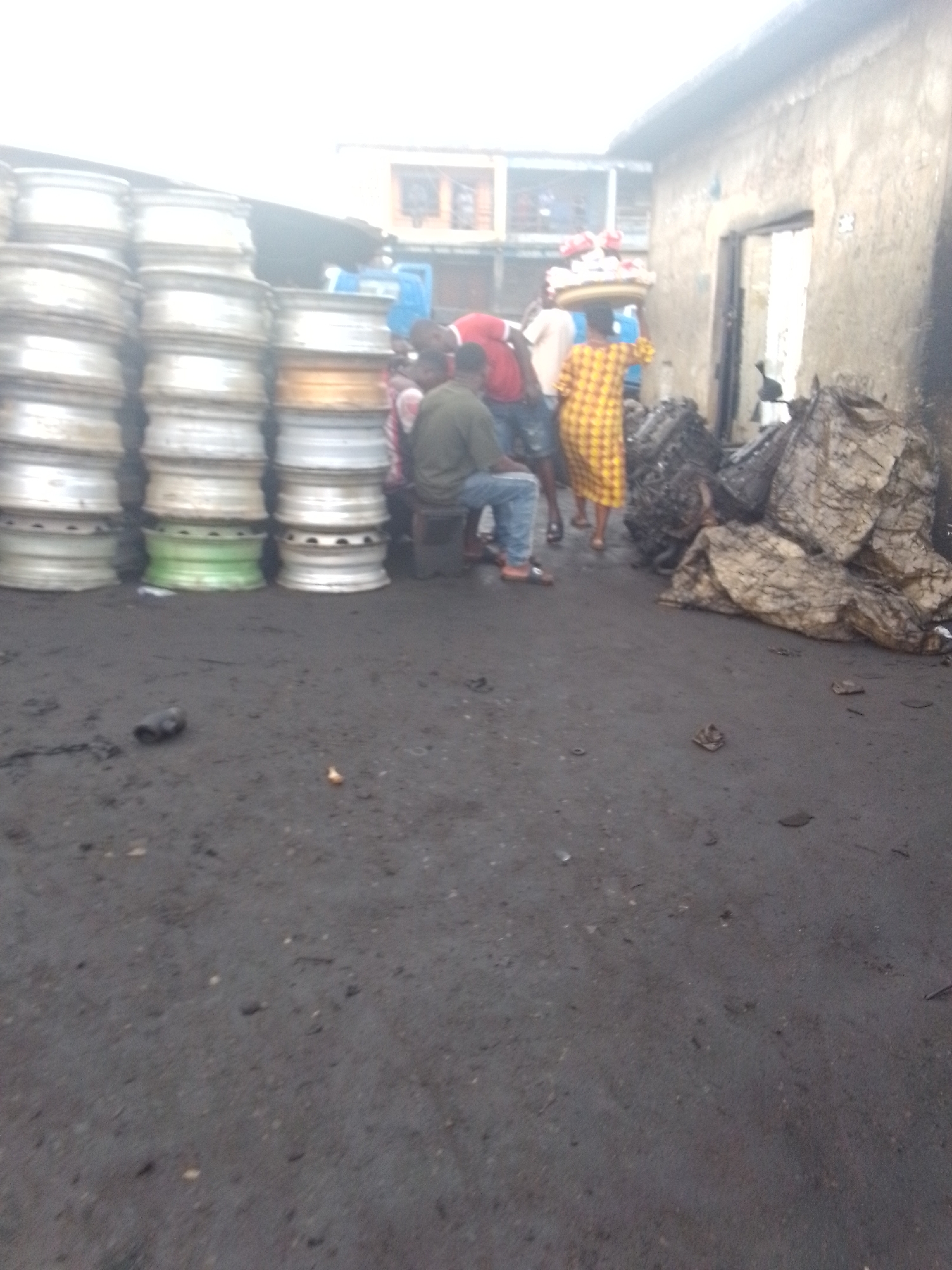The image shows a scene in what appears to be a marketplace or industrial area in Onitsha, Nigeria. The ground is dark and possibly oily. Stacks of metal rims are piled on the left side of the frame. A group of people are gathered near the rims and next to a building with an open doorway. One person is seated on a low stool, wearing a dark shirt and blue jeans. Another person wears a yellow patterned dress and carries something on their head. Bags or bundles of material are piled near the building. Other figures are present, with one wearing a red shirt.