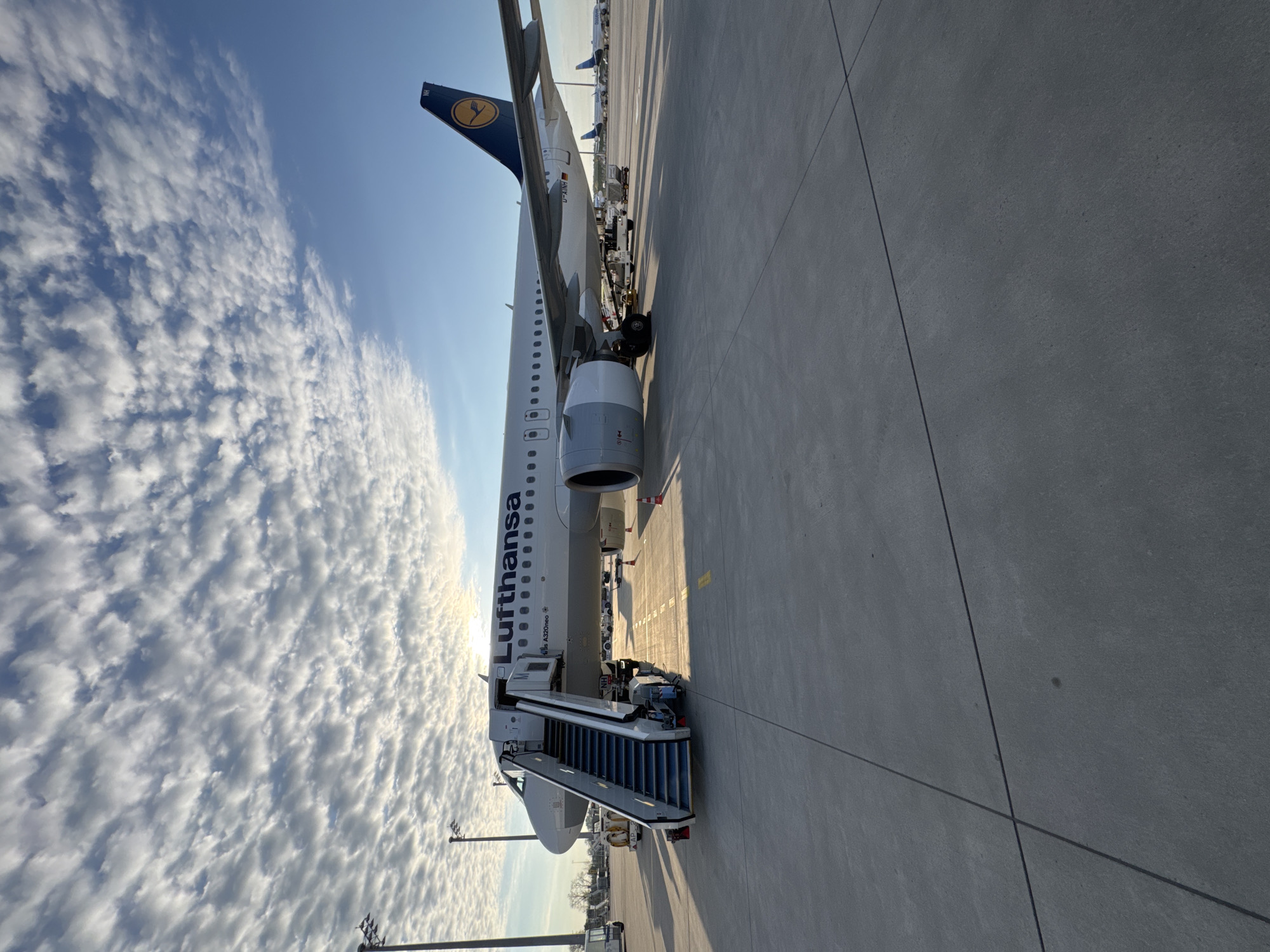 The image shows a Lufthansa Airbus A320neo aircraft on the tarmac at Munich Airport (MUC) in Oberding, Germany. The plane is connected to a set of mobile stairs, indicating passenger boarding or deboarding. The Lufthansa logo is visible on the plane's tail and fuselage. The sky is partly cloudy with a bright sun. The ground is concrete.