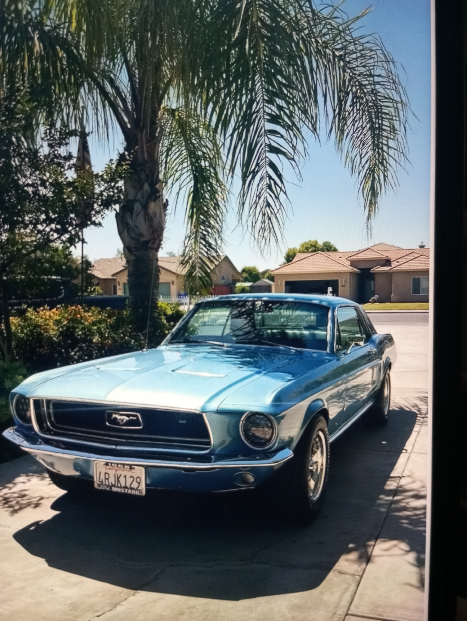 A light blue 1968 Ford Mustang is parked in a driveway. The car is a coupe and has a white stripe down the middle of the hood. The license plate reads "4RJK129" and has "1968 CALIFORNIA" and "FOR MUSTANG" printed on it. The car is parked in front of a house with a tile roof and a manicured lawn. A palm tree is visible in the background, casting shadows on the car. The sky is clear and blue. The location is a residential street in Zahorna, Moldova.