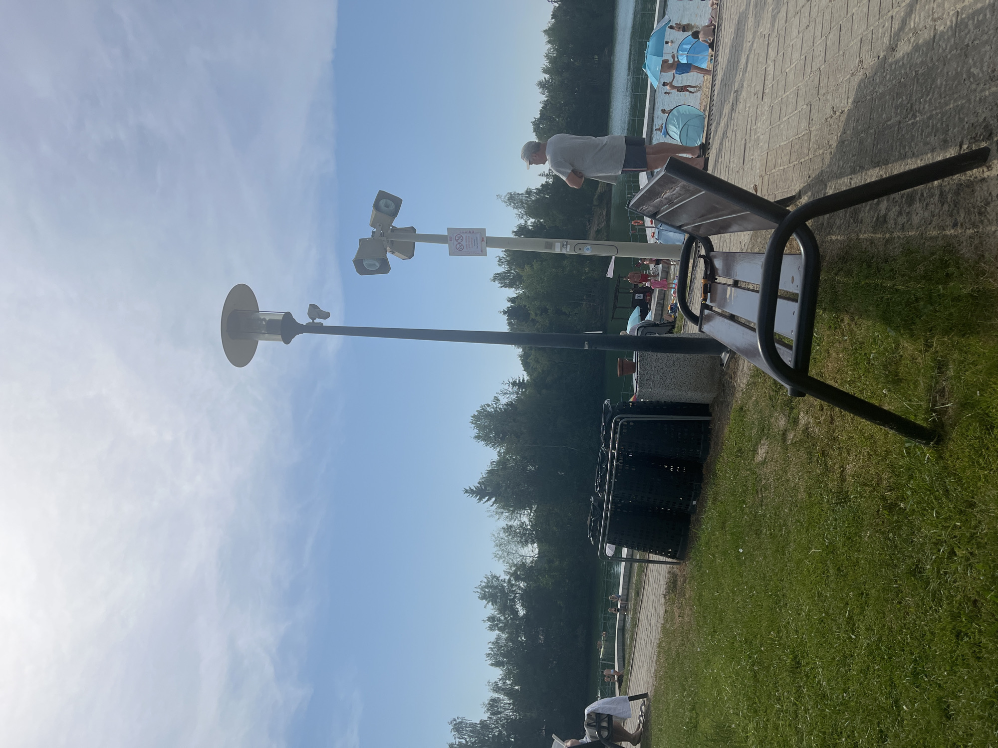 The image depicts a daytime scene at an outdoor recreational area, likely a beach or poolside, characterized by a bright, sunny sky with scattered clouds. In the foreground, a dark metal bench with wooden slats is positioned on a patch of green grass, casting a shadow. To the left of the bench is a lamppost with multiple lights. Behind the bench, a paved walkway runs alongside a body of water, with a treeline bordering the far bank.
Several people are visible in the background, dressed in summer attire. Some are gathered near the water's edge, while others are walking along the path or sitting on the beach. There are blue umbrellas and beach tents set up, suggesting a day of leisure. A man in a grey t-shirt and dark shorts stands near the lamppost, looking down. Two large, dark-colored trash receptacles are situated next to the walkway.
A sign is affixed to the lamppost, displaying two circular icons with red diagonal lines through them, indicating prohibitions. The overall atmosphere appears relaxed and summery. The time of day is suggested to be afternoon due to the presence of shadows and the bright sunlight. The setting is a public area designed for recreation, with elements such as water, seating, and amenities for waste disposal.