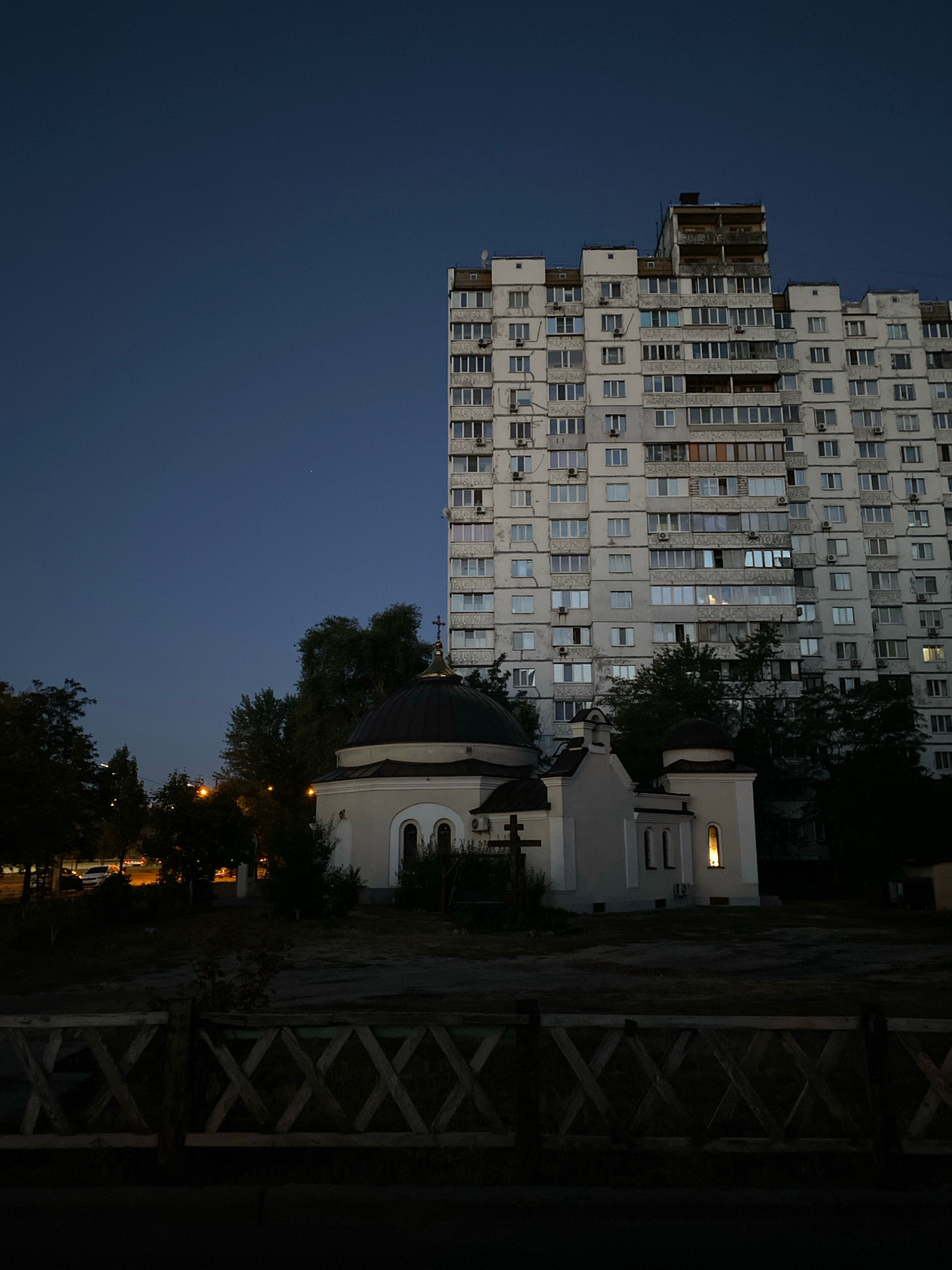 An exterior, medium shot captures a low-rise building with a domed roof, resembling a chapel, in the foreground, set against the backdrop of a tall, multi-story apartment building. The scene is illuminated by ambient light, suggesting it is either dusk or dawn. The sky is a deep, clear blue.
The chapel, painted in light beige, features a dark, rounded dome topped with a golden cross. It has arched windows and a smaller, square window on the right side of the main structure, which emits a warm, yellow light. A darker, smaller dome, also topped with a cross, is visible on the right side of the chapel. To the left of the chapel, a wooden cross stands upright.
The apartment building is a large, rectangular structure with numerous windows and air conditioning units visible on its facade. The windows are mostly dark, with some showing faint lights. The building's exterior appears to be weathered, with some visible signs of age.
In the background, trees are sparsely scattered, with some foliage visible between the chapel and the apartment building. A street with car lights is faintly visible to the left of the chapel. A wooden fence with a diamond pattern runs across the bottom of the frame, obscuring the lower portion of the scene. The overall impression is of an urban environment at night.