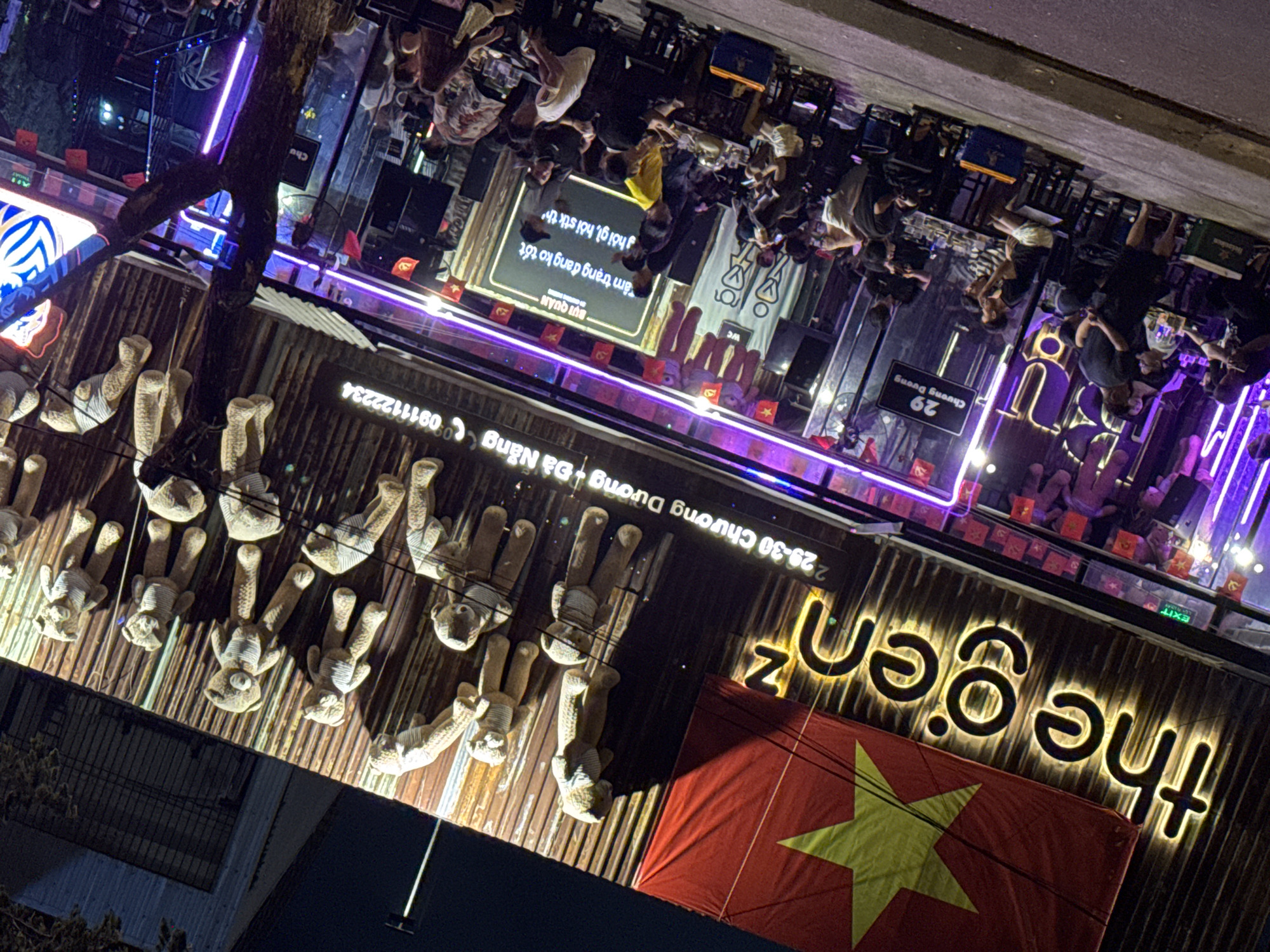 The image shows a street-level view of a busy establishment at night, likely a bar or restaurant, in Đà Nẵng, Vietnam. The facade of the building is adorned with a large Vietnamese flag and numerous plush teddy bears. A neon sign in English reads "the gen z." Below this, in smaller Vietnamese text, is an address: "29-30 Chuong Duong + Da Nang" and a phone number "0911122234".
The foreground is dominated by patrons sitting at tables on the sidewalk, engaged in conversation and consuming drinks. The atmosphere is lively, with purple neon lights illuminating the area. People are dressed casually, and some are clearly interacting with each other. The street is paved, and a few vehicles, possibly motorbikes, are visible in the background. The overall impression is one of a vibrant nightlife scene in a Vietnamese city.