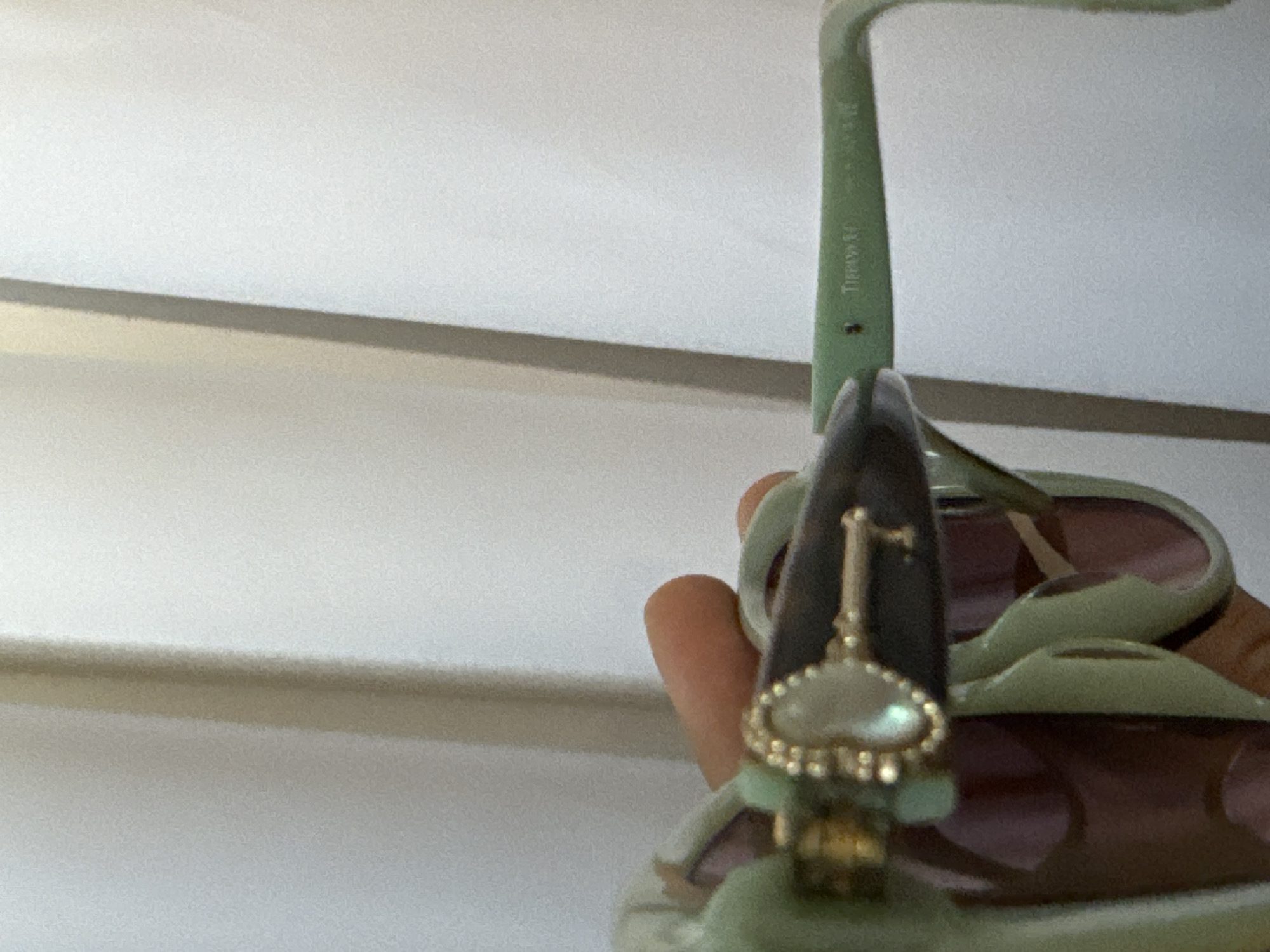 The image shows a pair of light green Tiffany & Co. sunglasses with brown lenses. A gold-colored key pendant is attached to the center of the sunglasses. The background is a white wall with a light brown trim. The photo was likely taken indoors. There is no indication of Davao City or other location. No people are visible in the image.