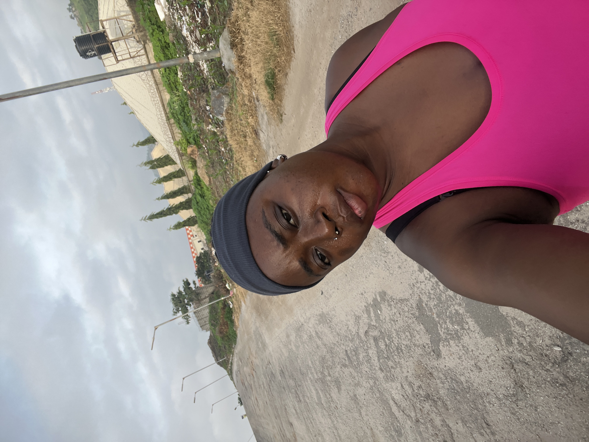 A selfie taken outdoors in Abuja, Nigeria shows a dark-skinned woman wearing a gray beanie and a bright pink tank top. She is looking at the camera with a neutral expression. The background is an outdoor scene with a dirt and concrete road leading uphill. There are streetlights visible along the road, and on the right side of the frame, there is a building with a patterned roof, a water tank, and some vegetation. The sky is overcast.