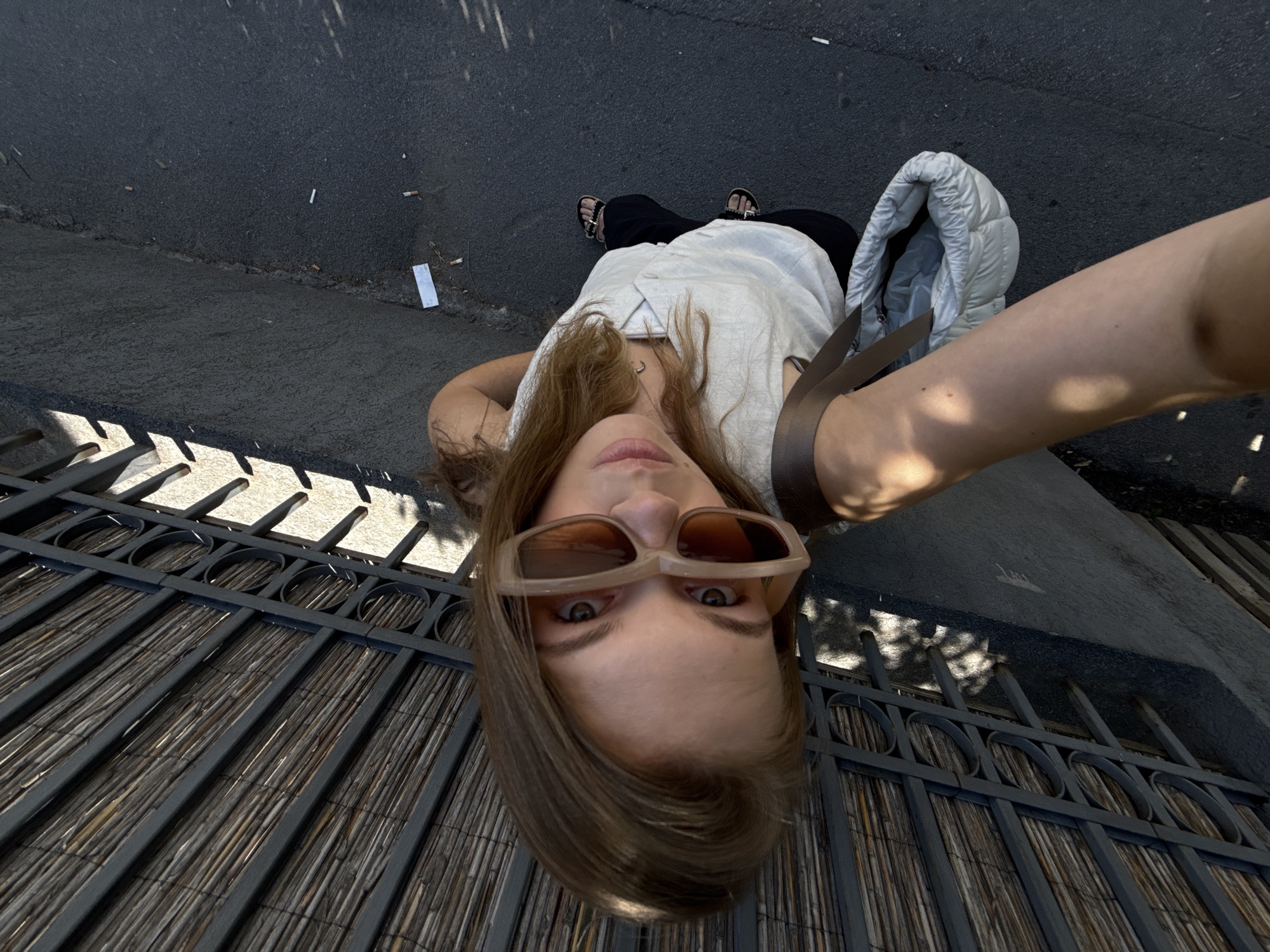 A young woman with long brown hair, wearing large, light-colored sunglasses, a white sleeveless top, black pants, and sandals, takes a high-angle selfie while standing on a paved area. She carries a light-colored puffy bag over her shoulder. Behind her, a dark metal fence with horizontal bamboo-like reeds forms a canopy overhead. Shadows from the fence and canopy are cast on the ground. The ground surface appears to be asphalt, with a slight incline. Several discarded cigarette butts are visible on the asphalt. The scene suggests an outdoor urban environment.