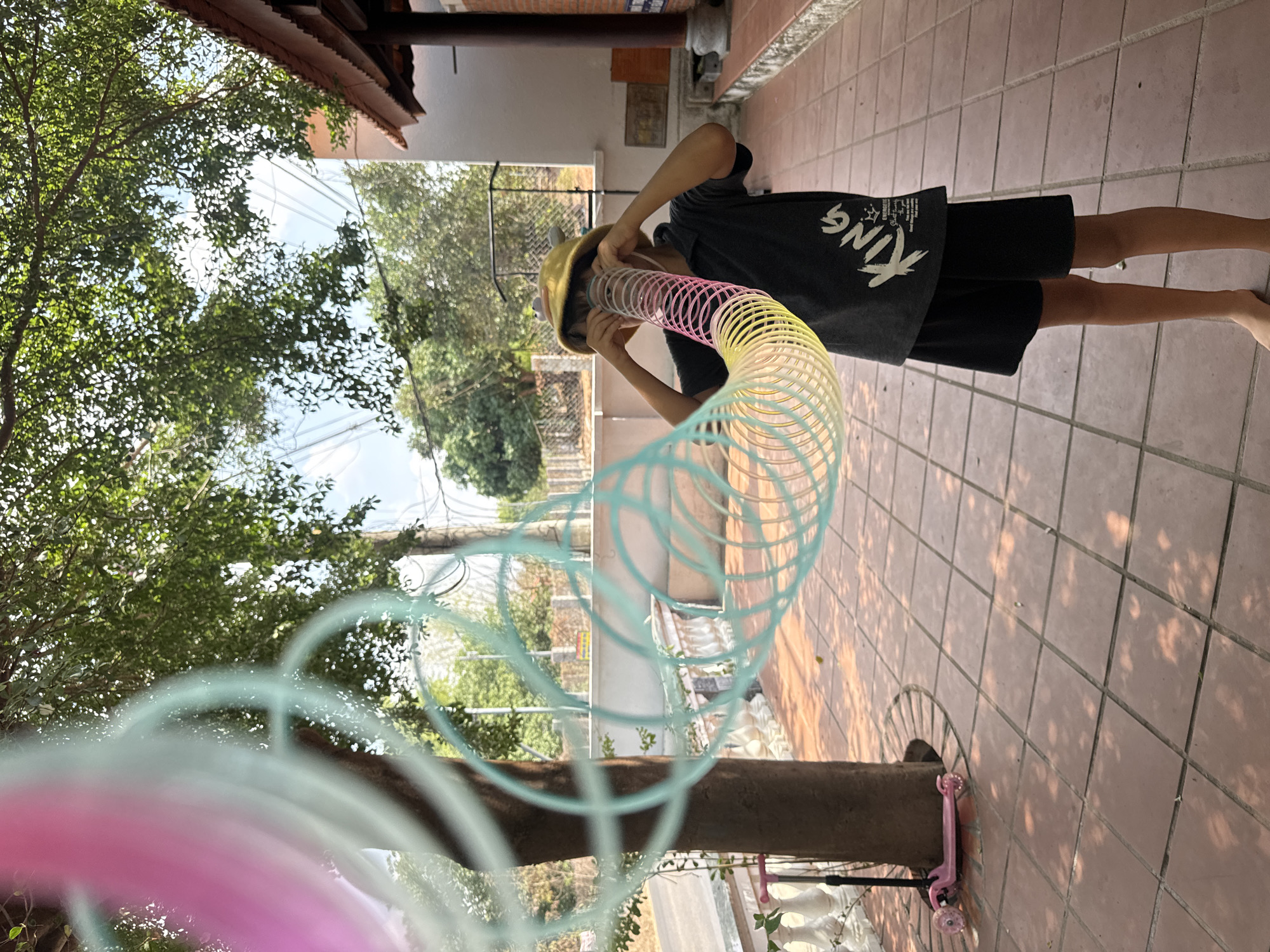 A child stands on a tiled patio in Mũi Né, Vietnam. The child, wearing a dark gray t-shirt with white lettering and black shorts, holds a colorful plastic spring toy up to their face, looking through it. The patio is composed of square, terracotta-colored tiles. Behind the child is a building with a roof overhang. To the left, a tree with green leaves fills a significant portion of the frame. A pink scooter sits near a support post on the right.