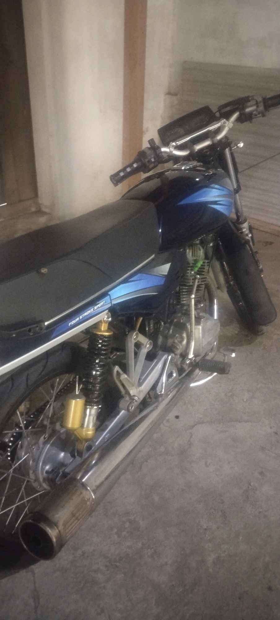 A side view of a dark blue and silver motorcycle parked on a concrete floor. The motorcycle has a black seat, a visible engine, a chrome exhaust pipe, and a spoked rear wheel with a gold shock absorber. A sticker on the side of the motorcycle reads "FOUR STROKE SPORT". The background consists of a textured wall and a wooden doorframe, suggesting an indoor or covered area. There are no people or active events depicted in the image. The scene is static and focused on the motorcycle itself.